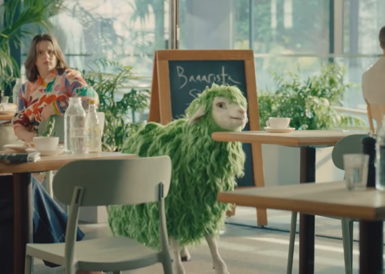 Ein grünes Schaf steht in einem modernen Café, eine Frau schaut überrascht dorthin.