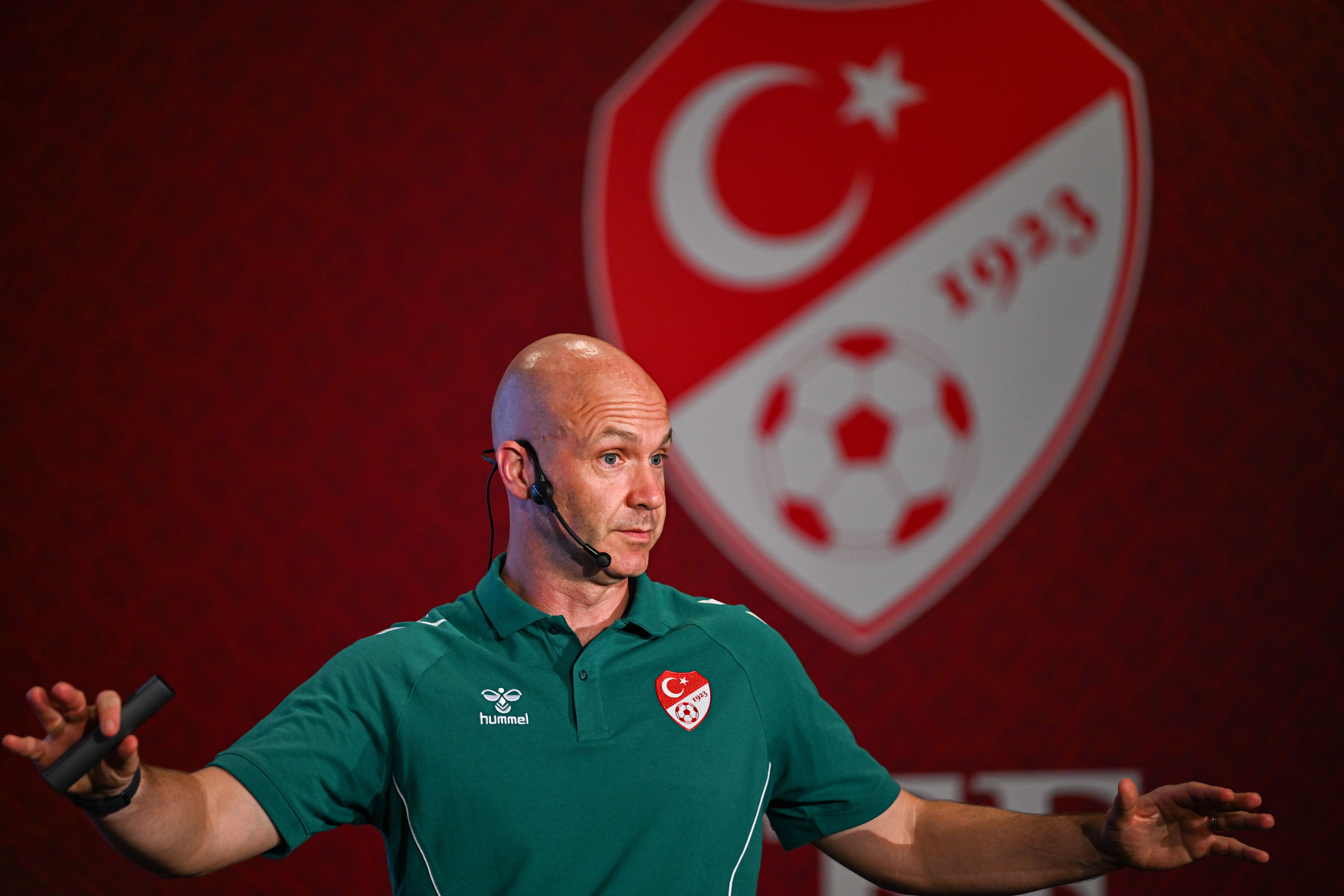 Anthony Taylor steht in einem grünen Polo-Shirt der TFF mit Ansteckmikrofon vor einer roten Wand mit dem Logo des türkischen Fußballverbands und gestikuliert mit den Händen.