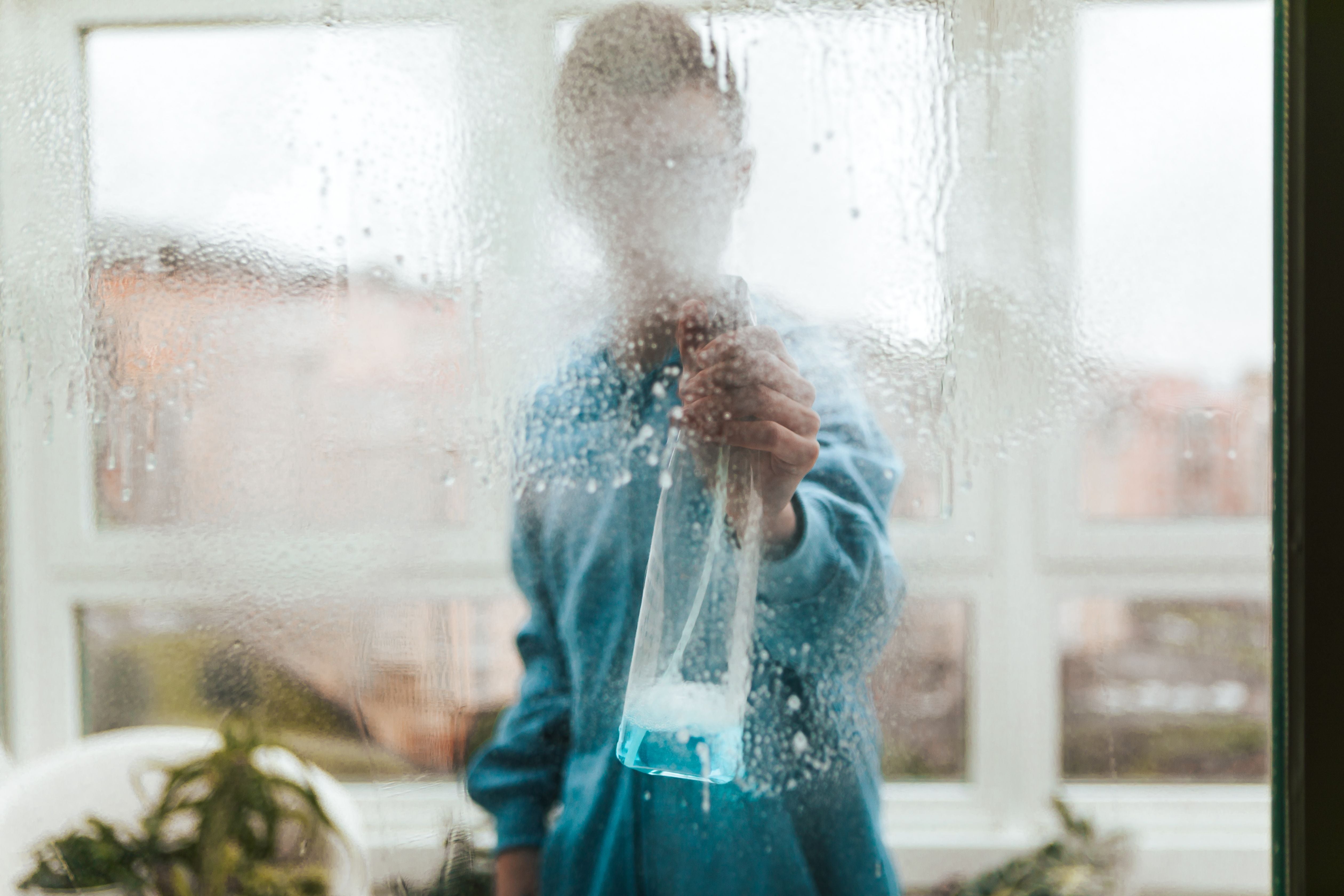Person sprüht Fensterreiniger auf eine Glasscheibe und putzt das Fenster von innen.