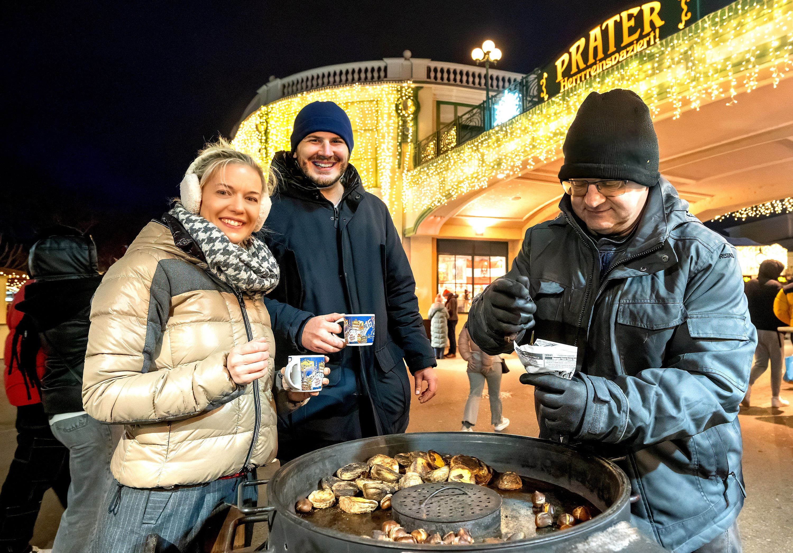 Drei Menschen genießen heiße Maroni und Glühwein am beleuchteten Wintermarkt Prater.