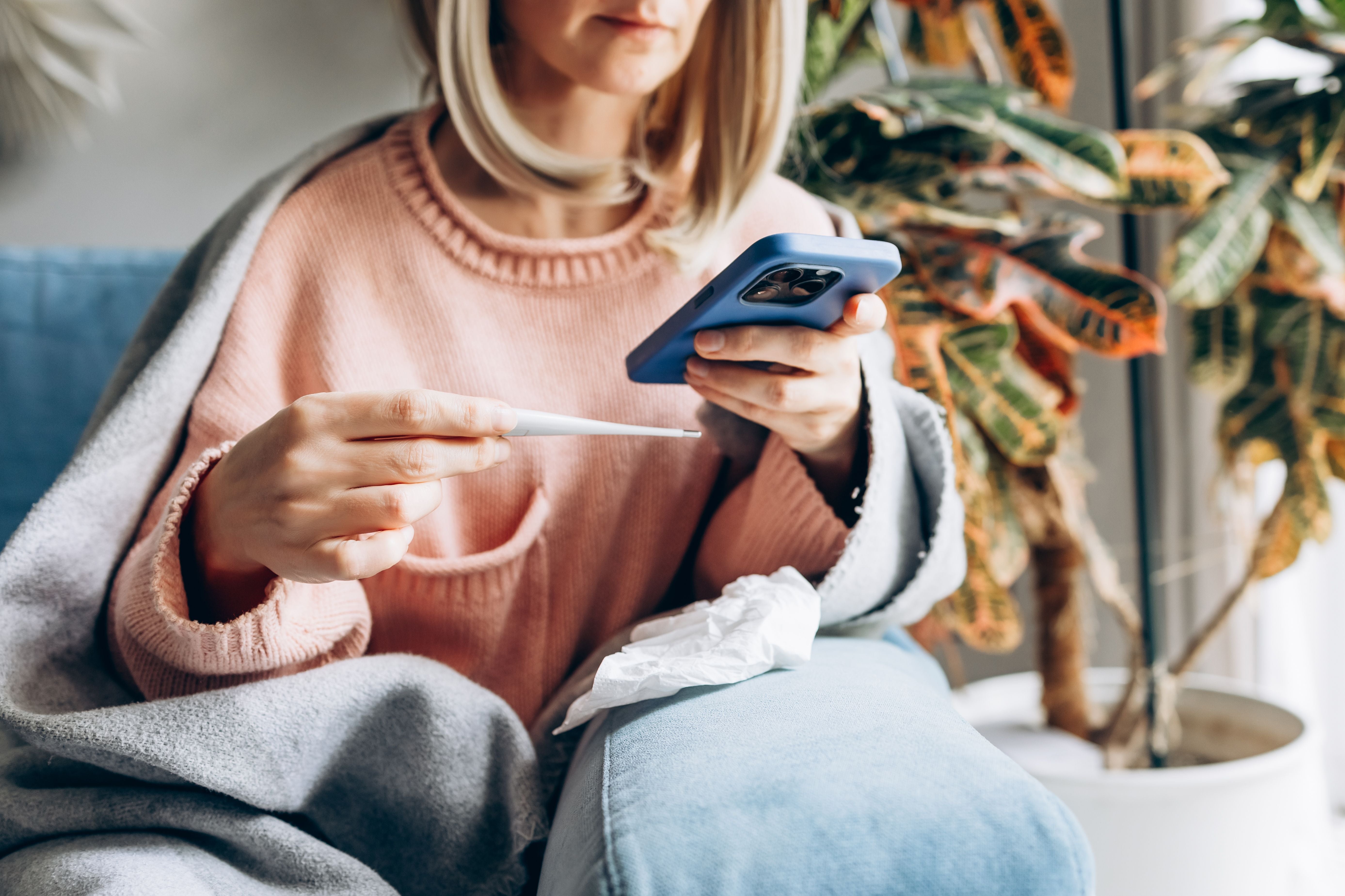 Frau mit Thermometer und Handy sitzt krank unter einer Decke auf dem Sofa.
