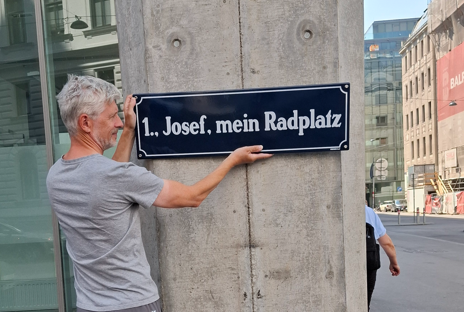 Ein Mann hält ein Schild mit der Aufschrift „1., Josef, mein Radplatz“ an eine Betonsäule.