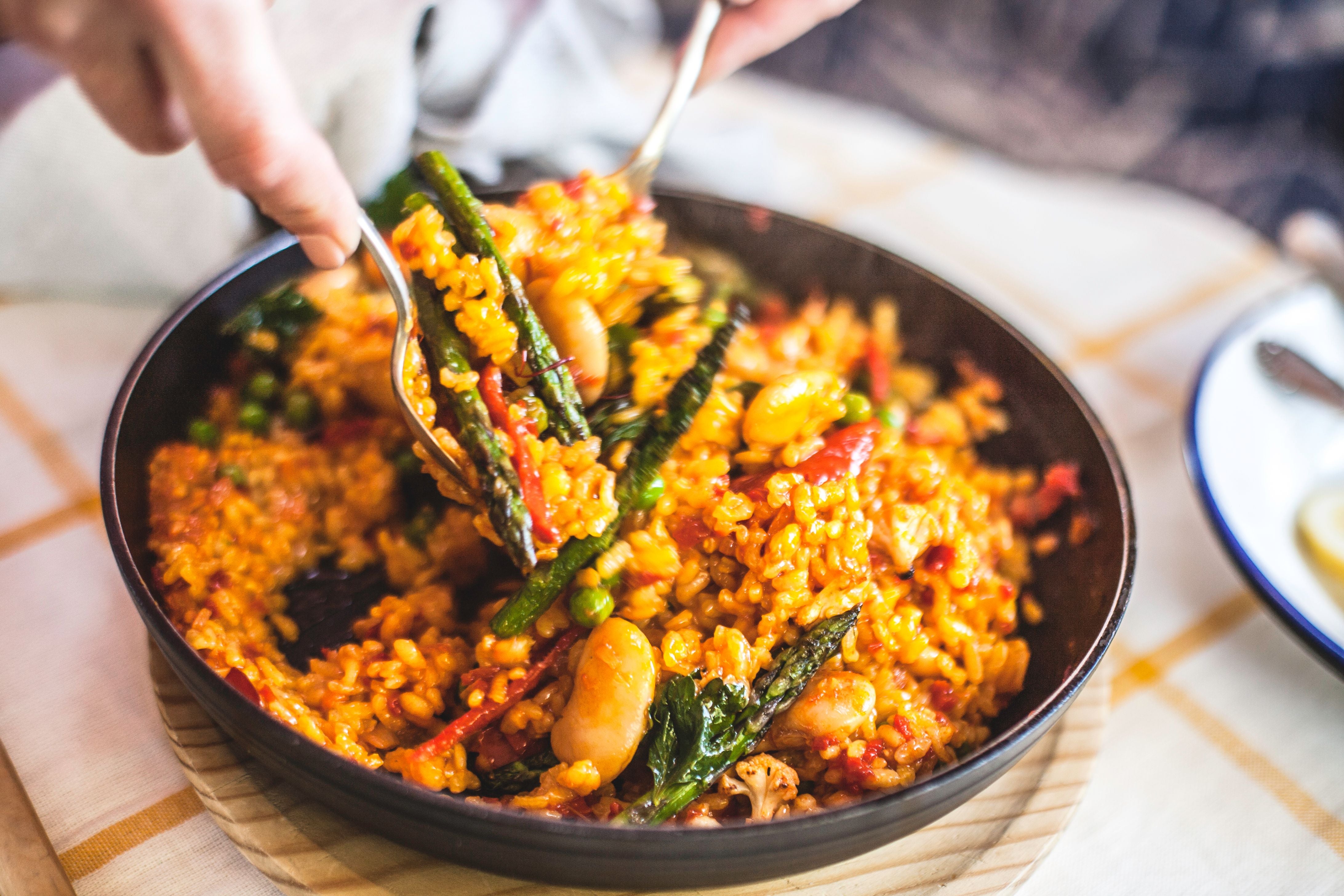 Eine Pfanne mit bunter Gemüsepaella wird auf einem gelb karierten Tischtuch serviert.