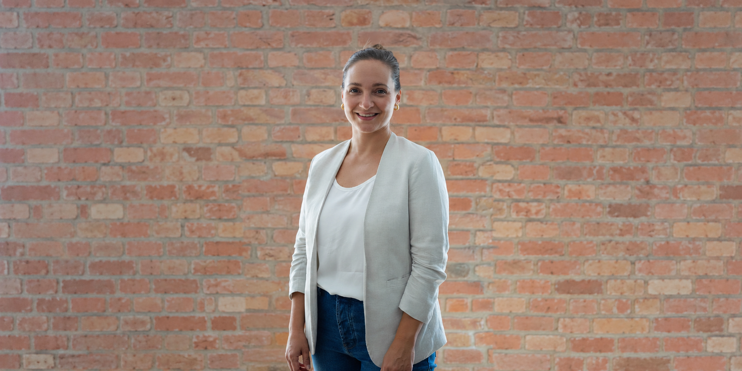 Frau steht lächelnd vor einer Backsteinwand und trägt eine helle Jacke und Jeans.