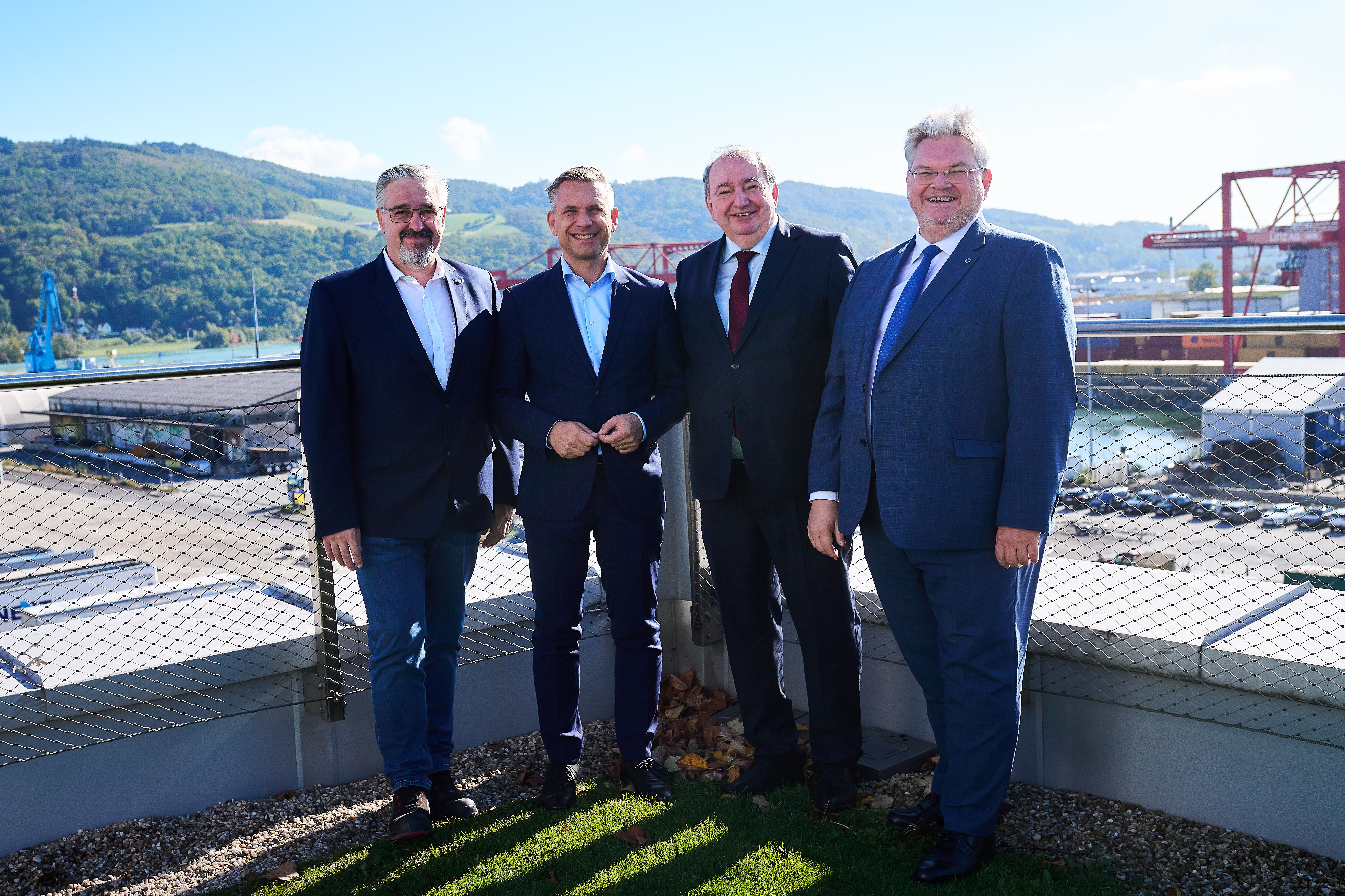 Vier Männer im Anzug stehen lächelnd auf einer Terrasse vor einem Industriehafen.