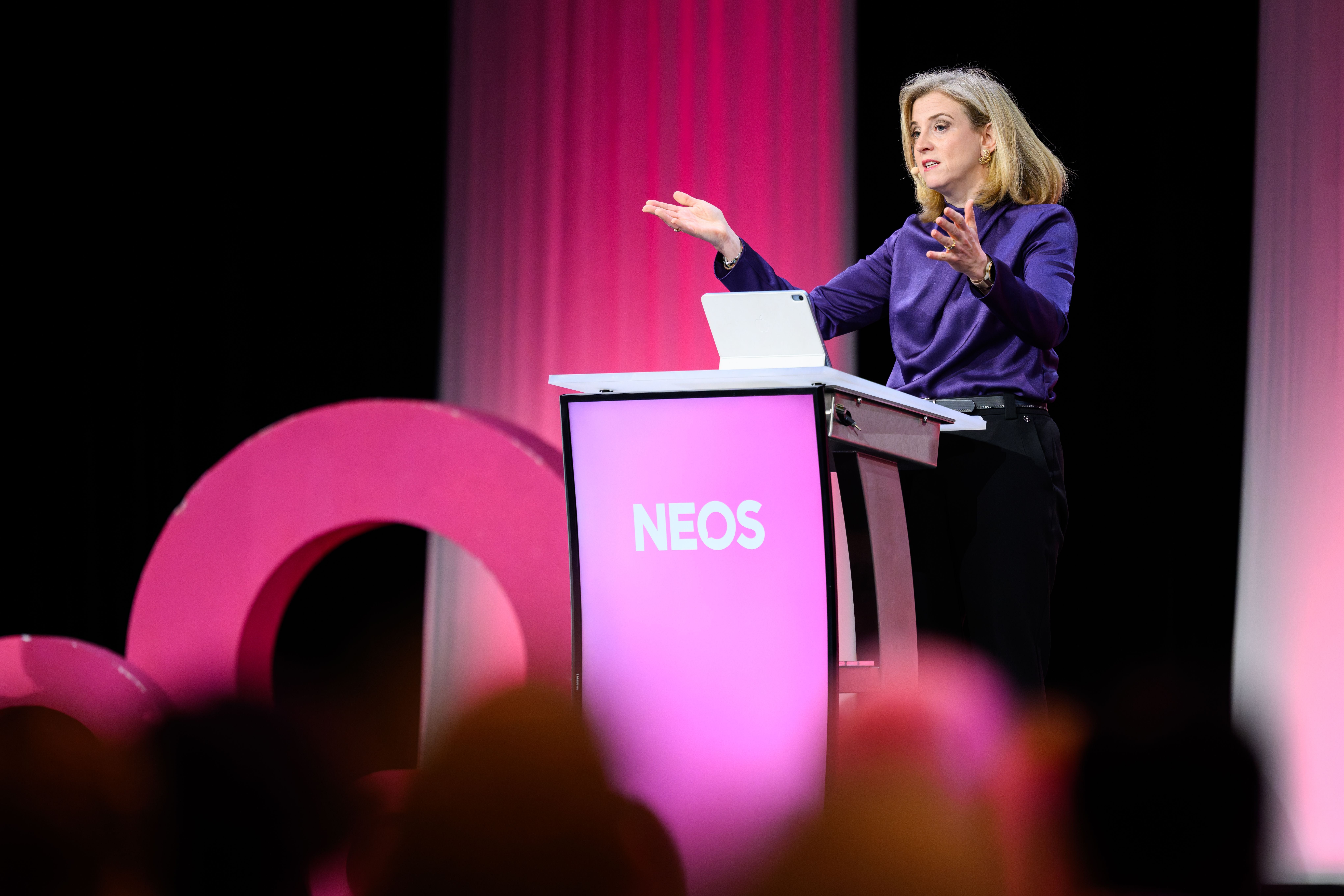 Frau am Rednerpult mit NEOS-Logo spricht bei einer Veranstaltung, Publikum im Vordergrund.