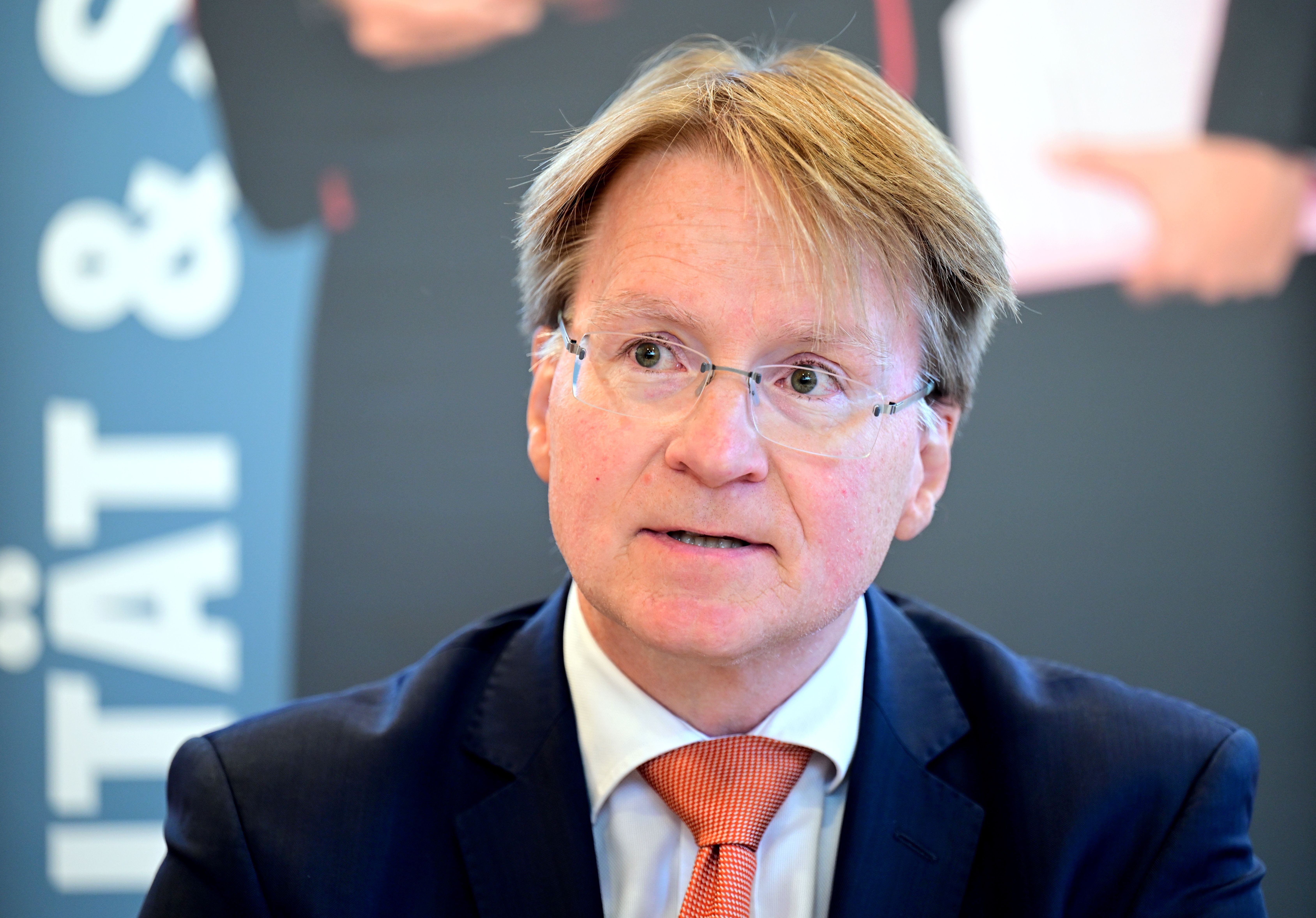 Ein Mann im Anzug mit Brille spricht bei einer Pressekonferenz in einem Innenraum.