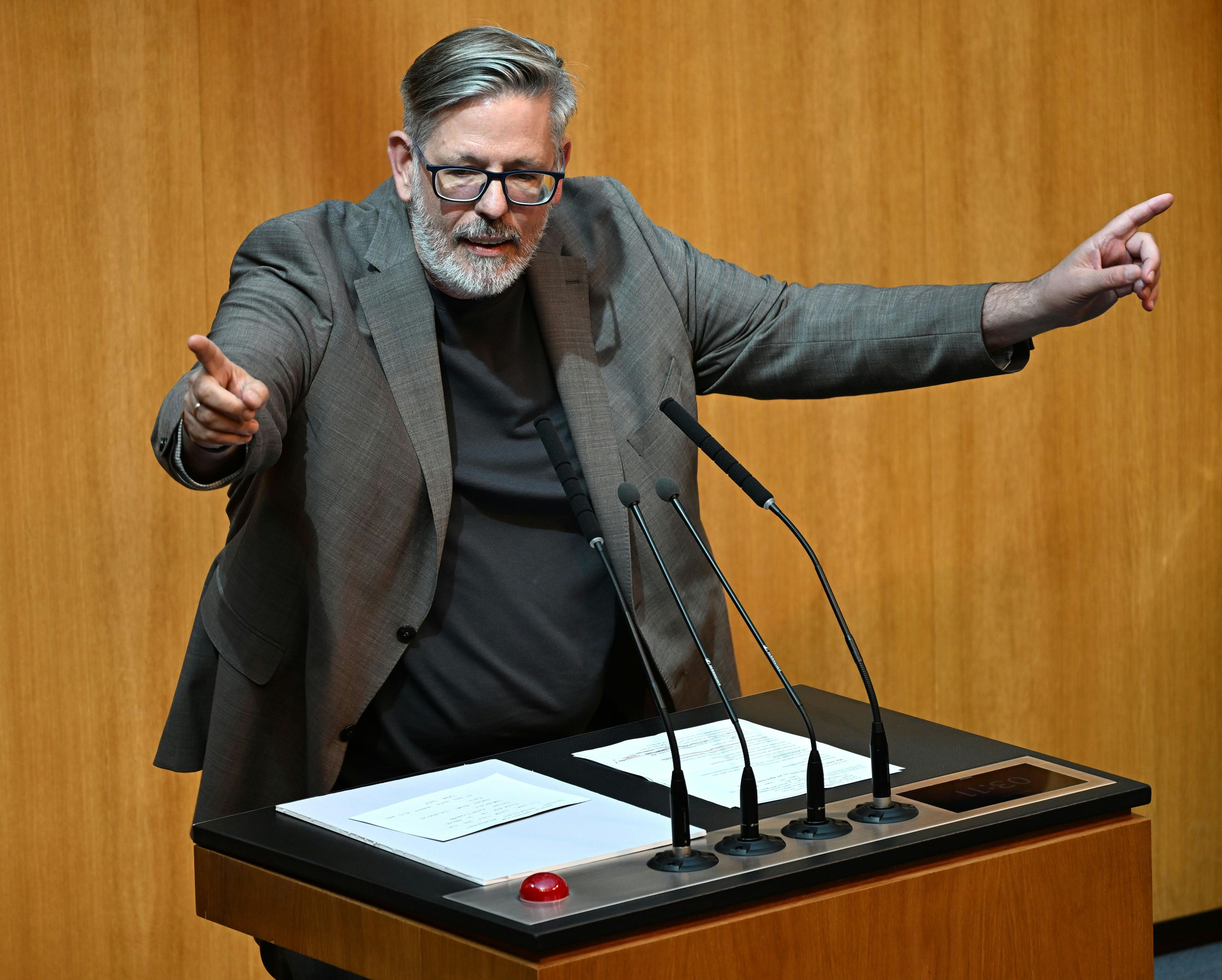 Mann spricht gestikulierend am Rednerpult im Parlament vor Mikrofonen und Papieren.