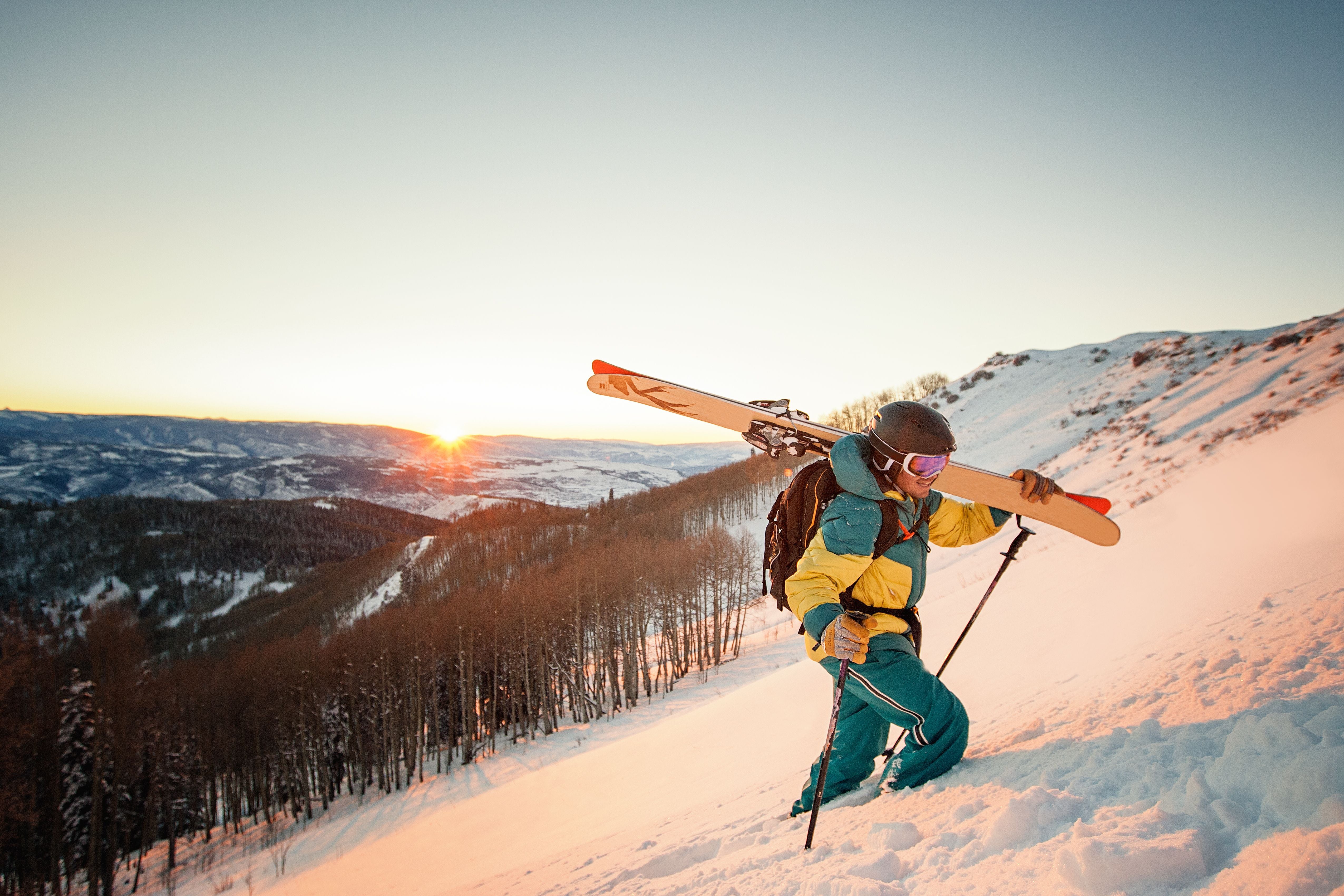 Skifahrer steigt mit Ausrüstung im Sonnenuntergang einen verschneiten Hang hinauf.