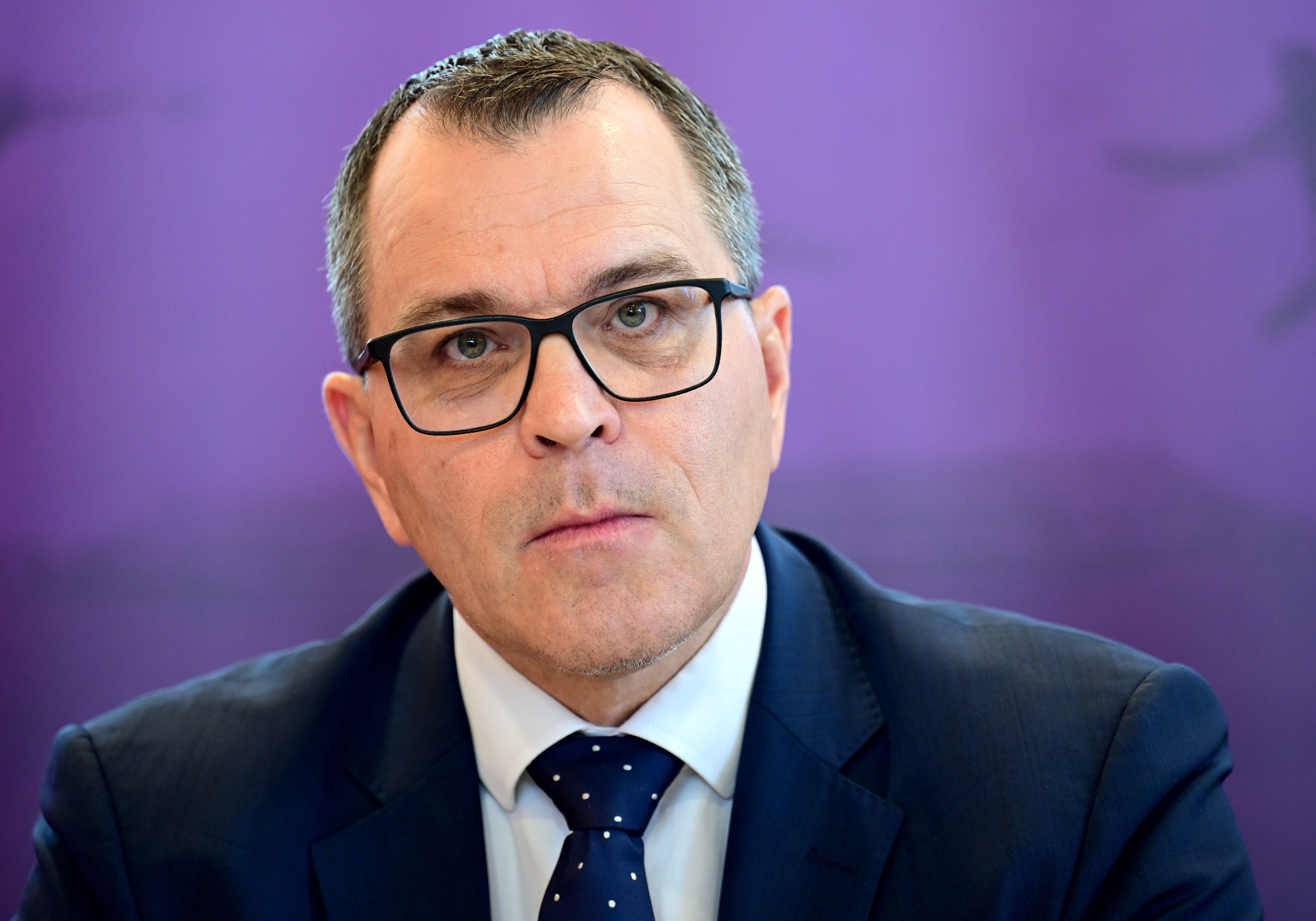 Mann mit Brille und Anzug vor lila Hintergrund bei einer Pressekonferenz.