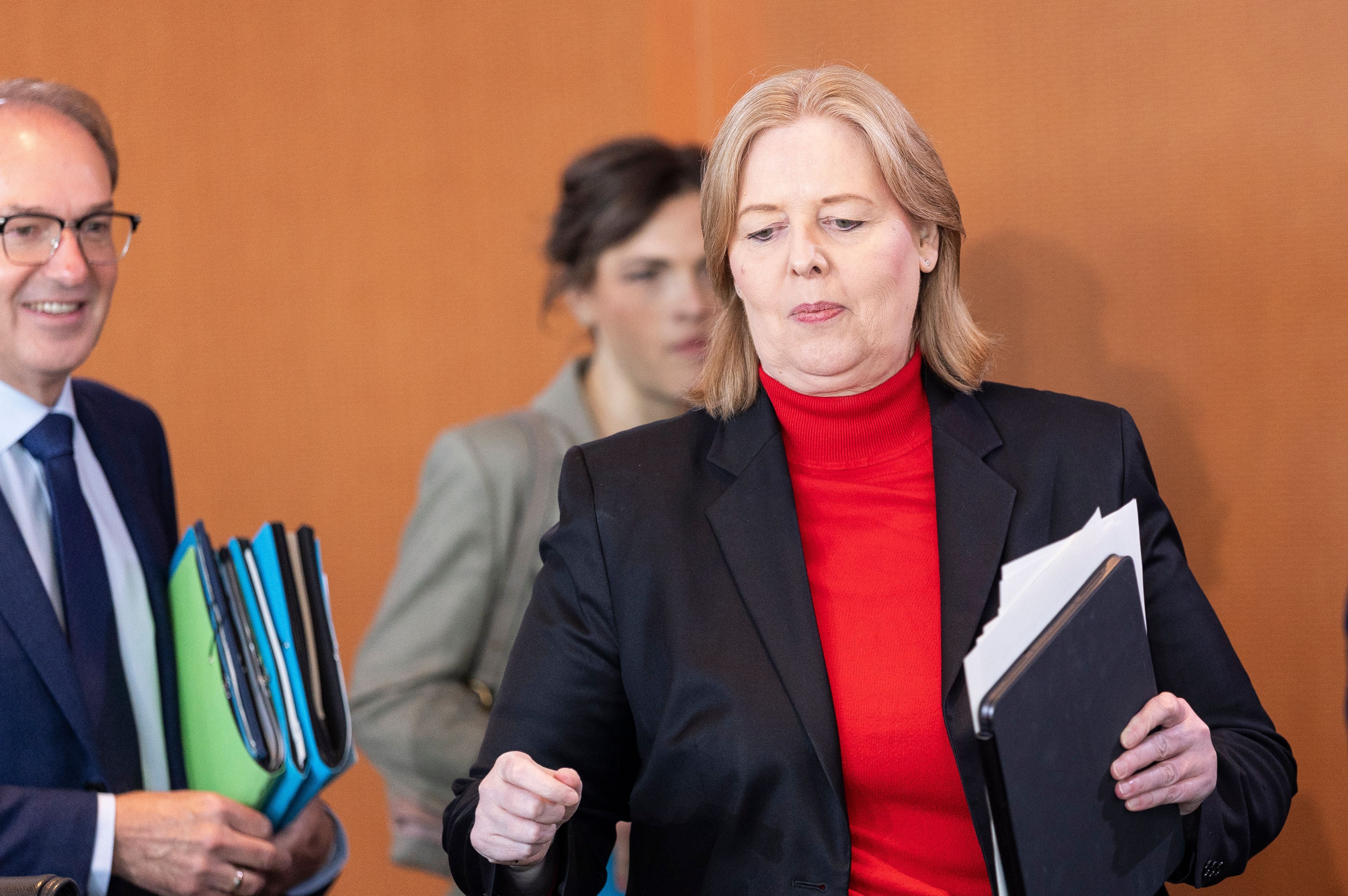 Eine Frau im schwarzen Blazer und rotem Rollkragen hält Akten und geht durch einen Raum.