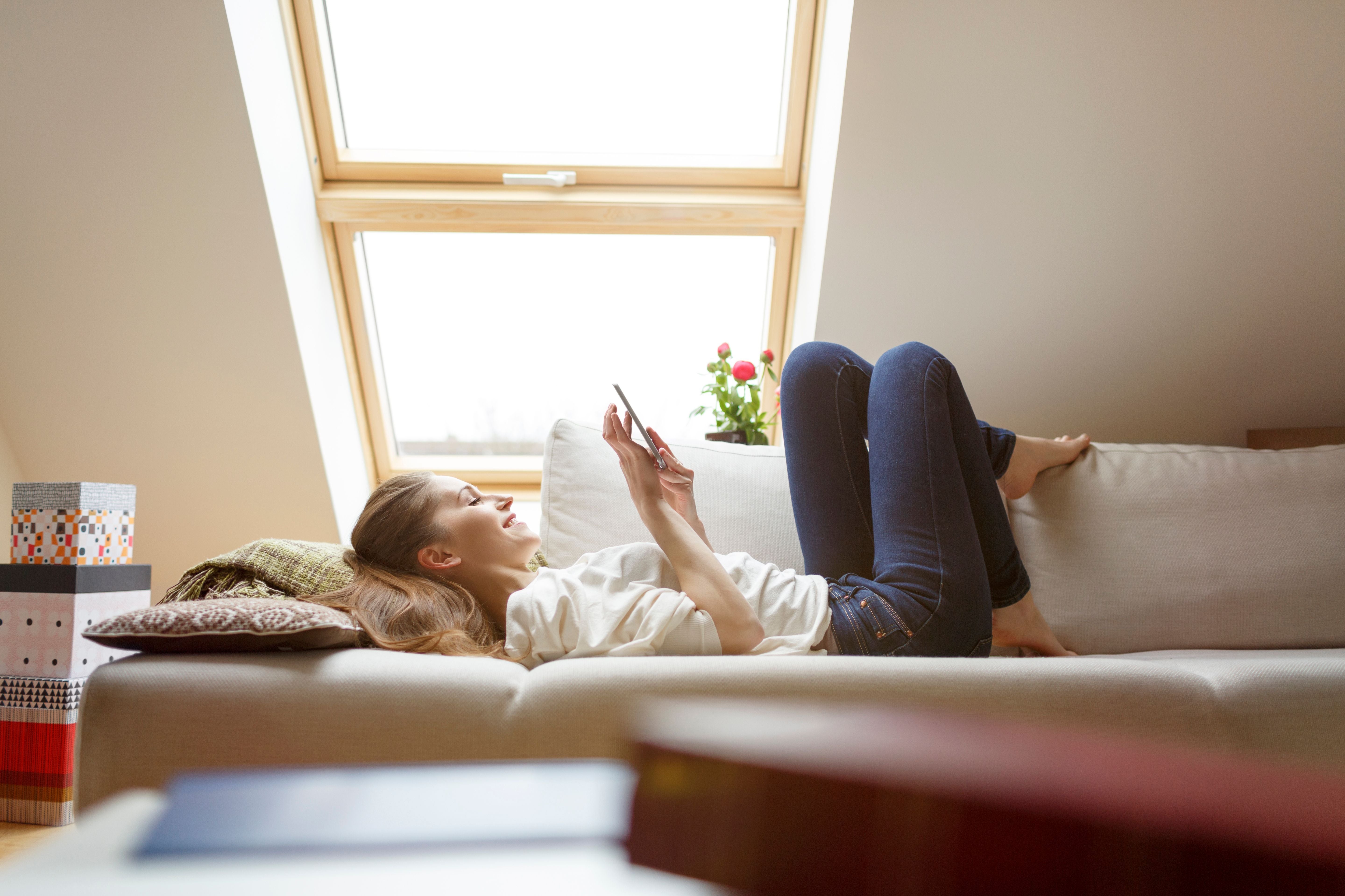 Junge Frau liegt entspannt auf dem Sofa unter einem Dachfenster und nutzt ein Smartphone.