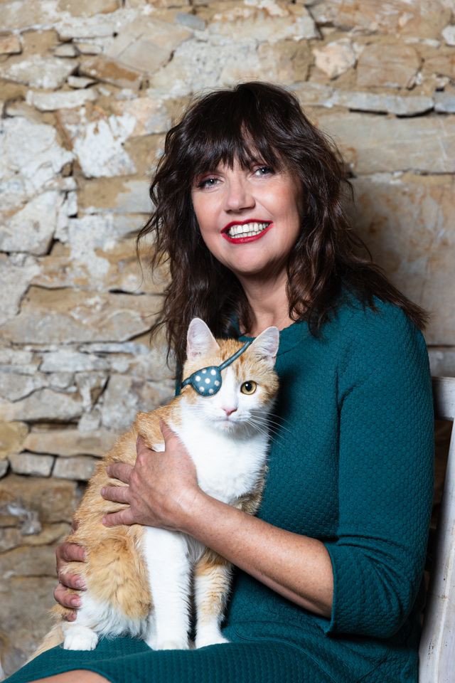 Martina Parker hält eine Katze mit Augenklappe auf dem Schoß, vor Steinmauer.