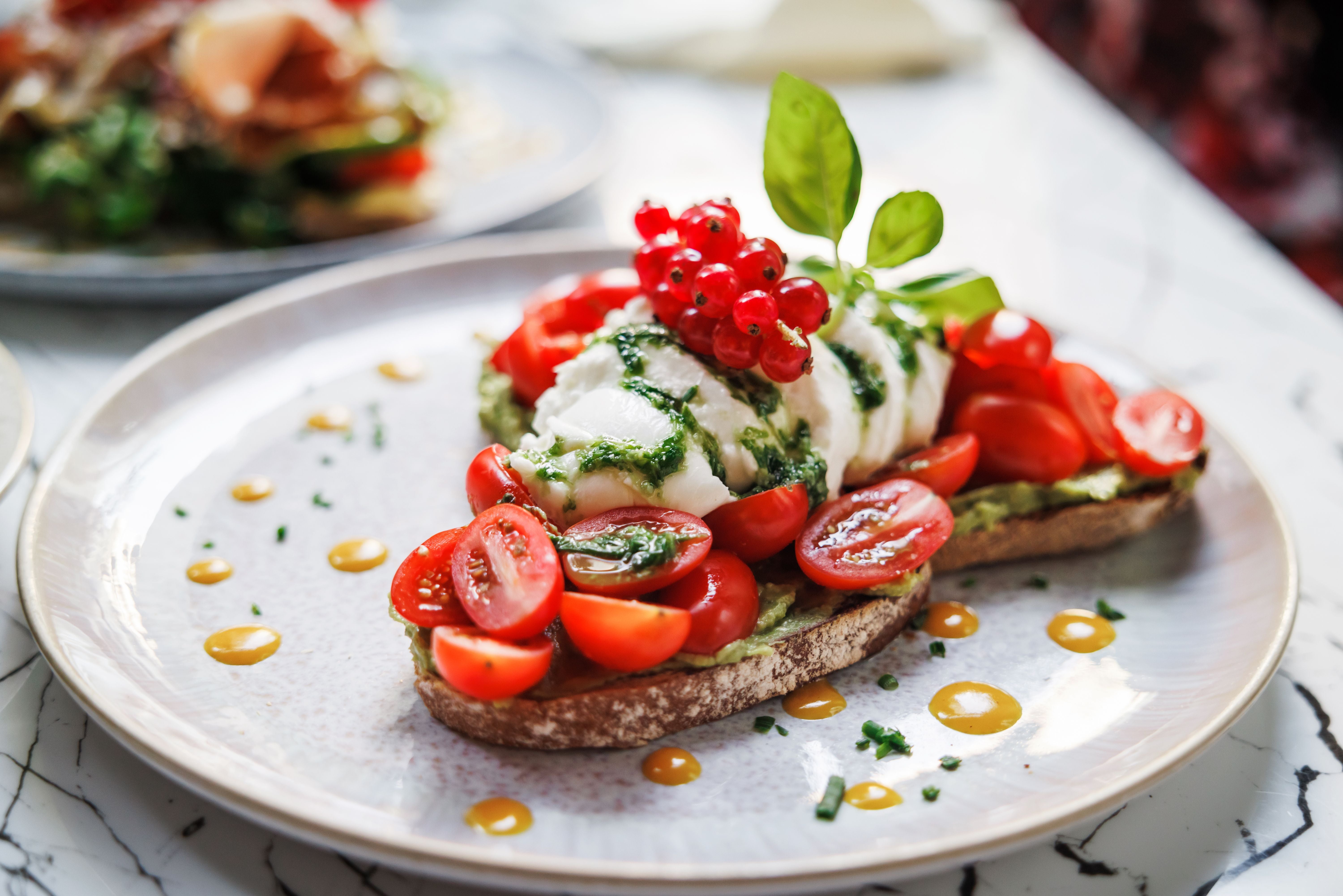 Avocado-Toast mit Kirschtomaten, Mozzarella, roten Johannisbeeren und Basilikum auf einem Teller.