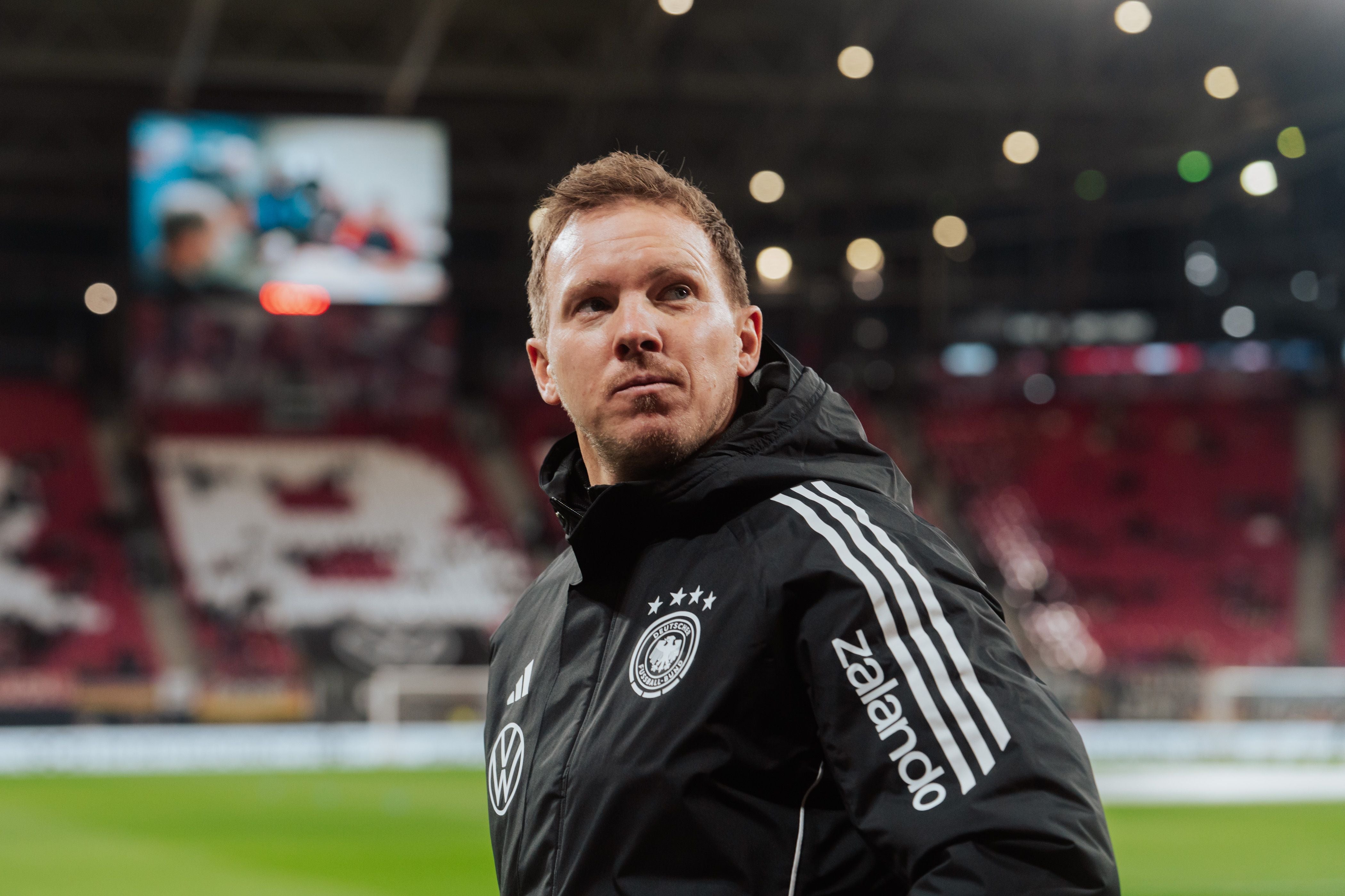Ein Fußballtrainer im Deutschland-Trainingsanzug blickt konzentriert im Stadion nach oben.