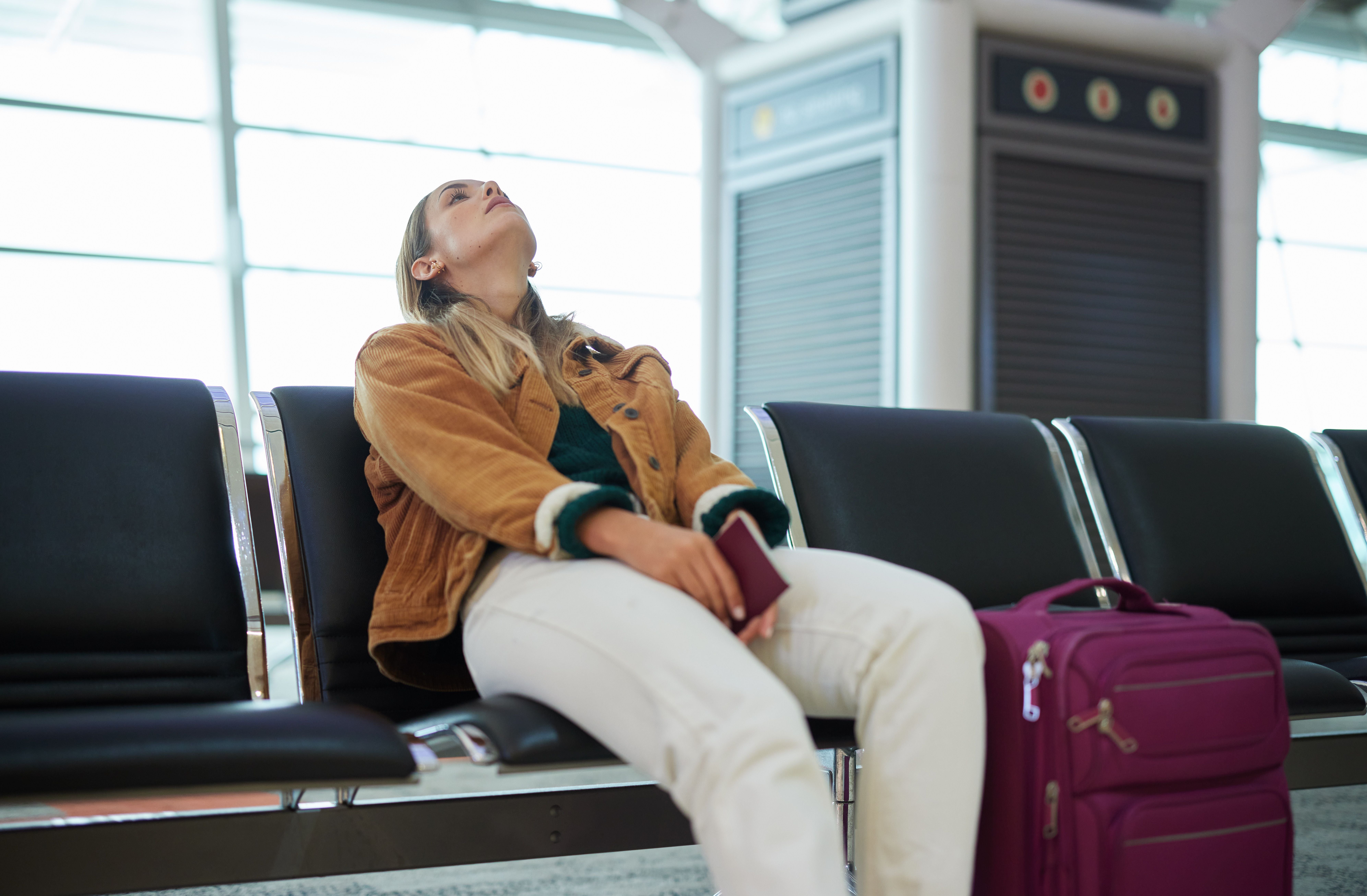 Frau sitzt gelangweilt am Flughafen mit Koffer und Pass und wartet auf ihren Flug.
