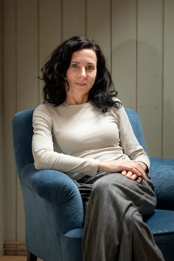 Frau mit dunklen Locken sitzt entspannt in einem blauen Sessel und blickt in die Kamera.