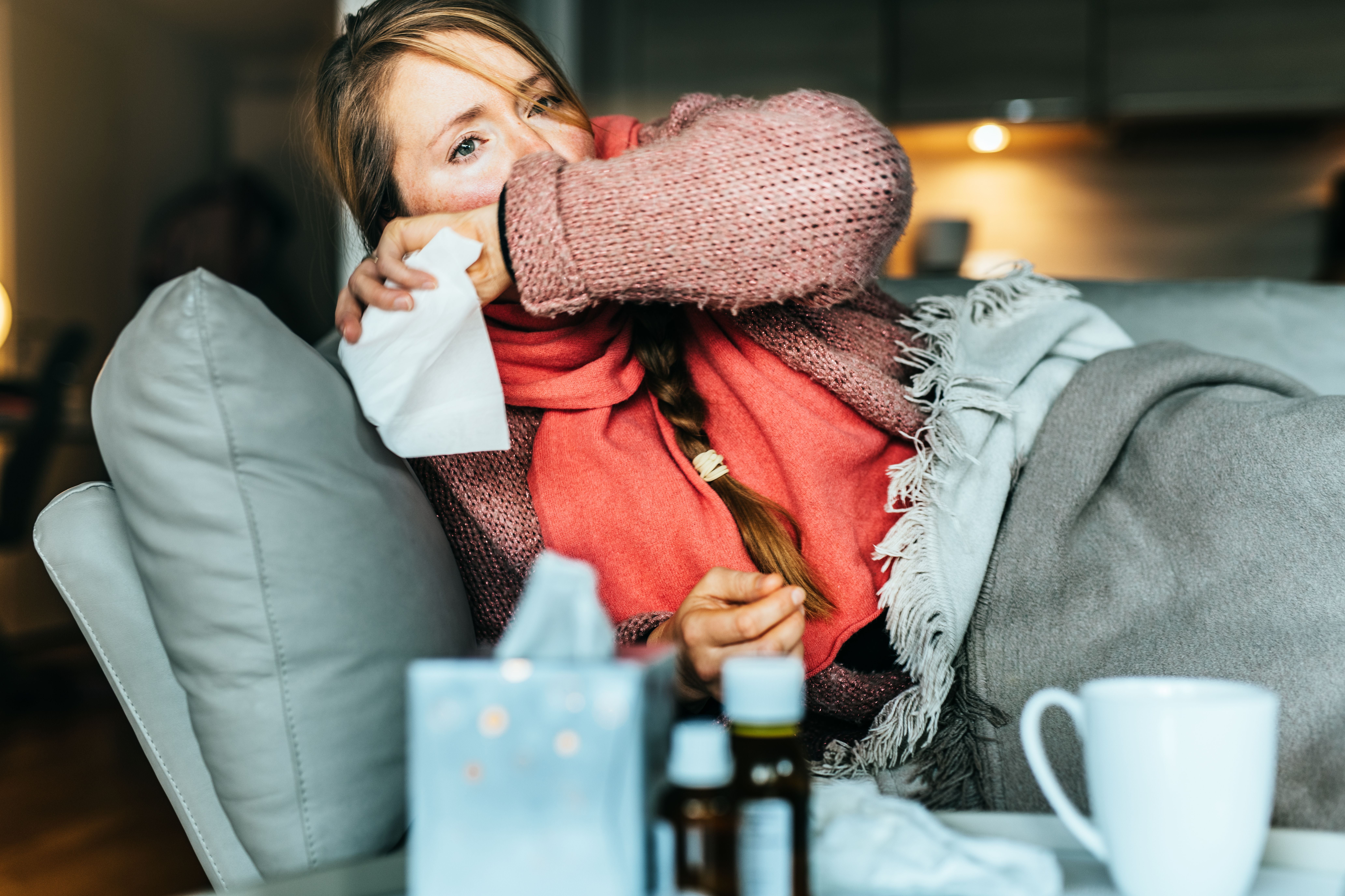 Frau liegt krank mit Schal und Decke auf dem Sofa, Medikamente und Tasse stehen auf dem Tisch.