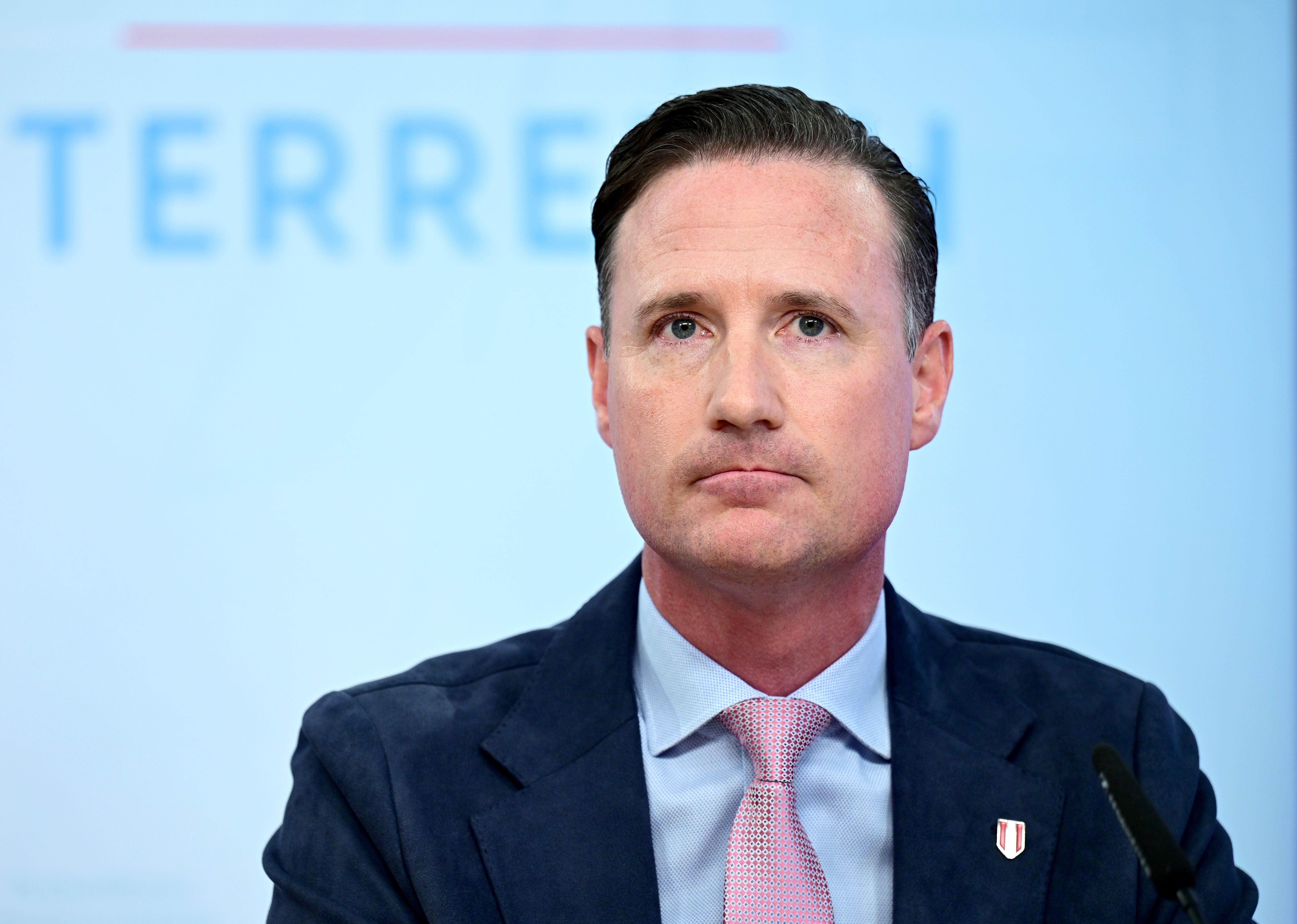 Ein Mann im Anzug und mit Krawatte sitzt vor einem Mikrofon bei einer Pressekonferenz.