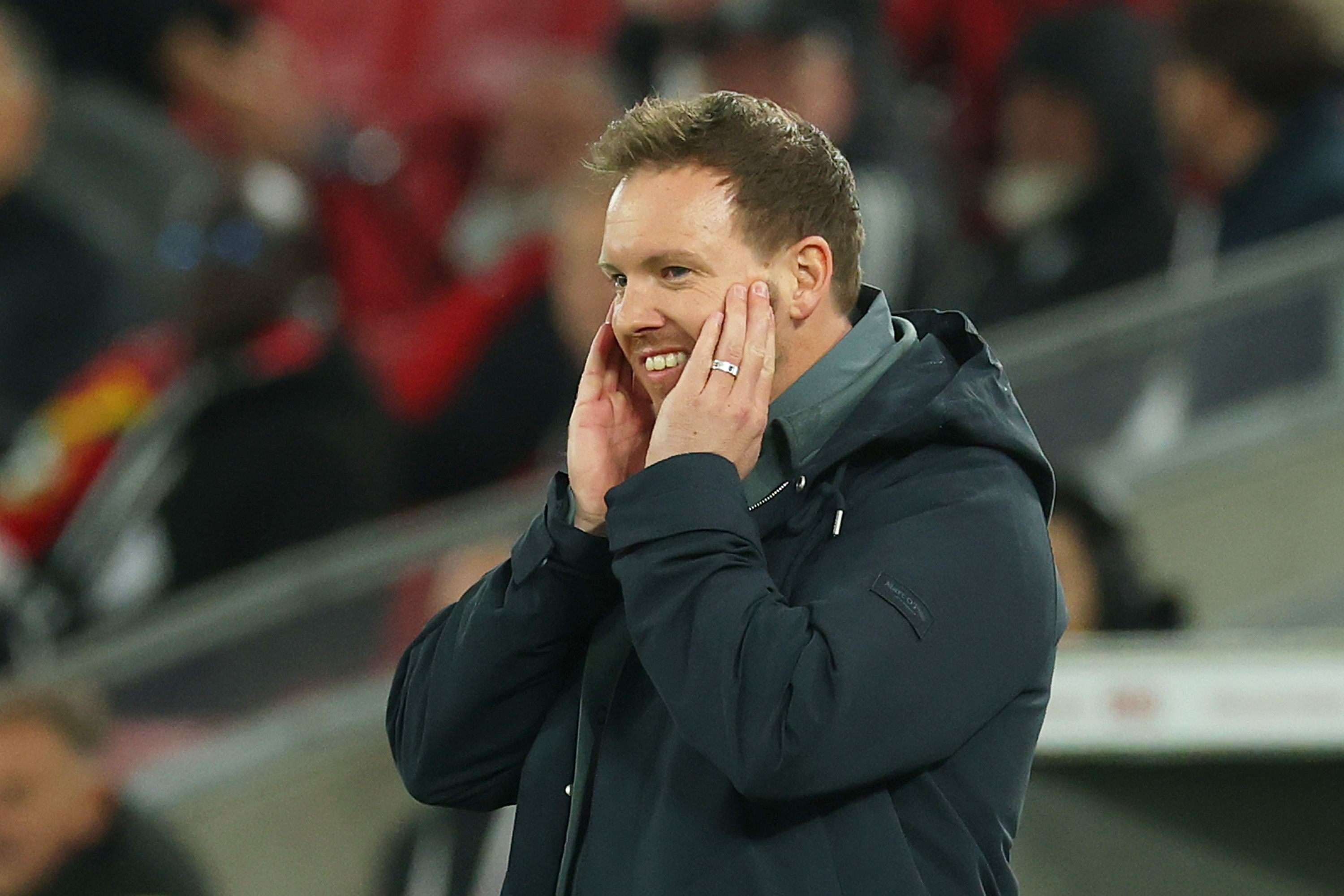 Ein Mann im dunklen Mantel hält sich lächelnd die Hände an die Wangen im Stadion.