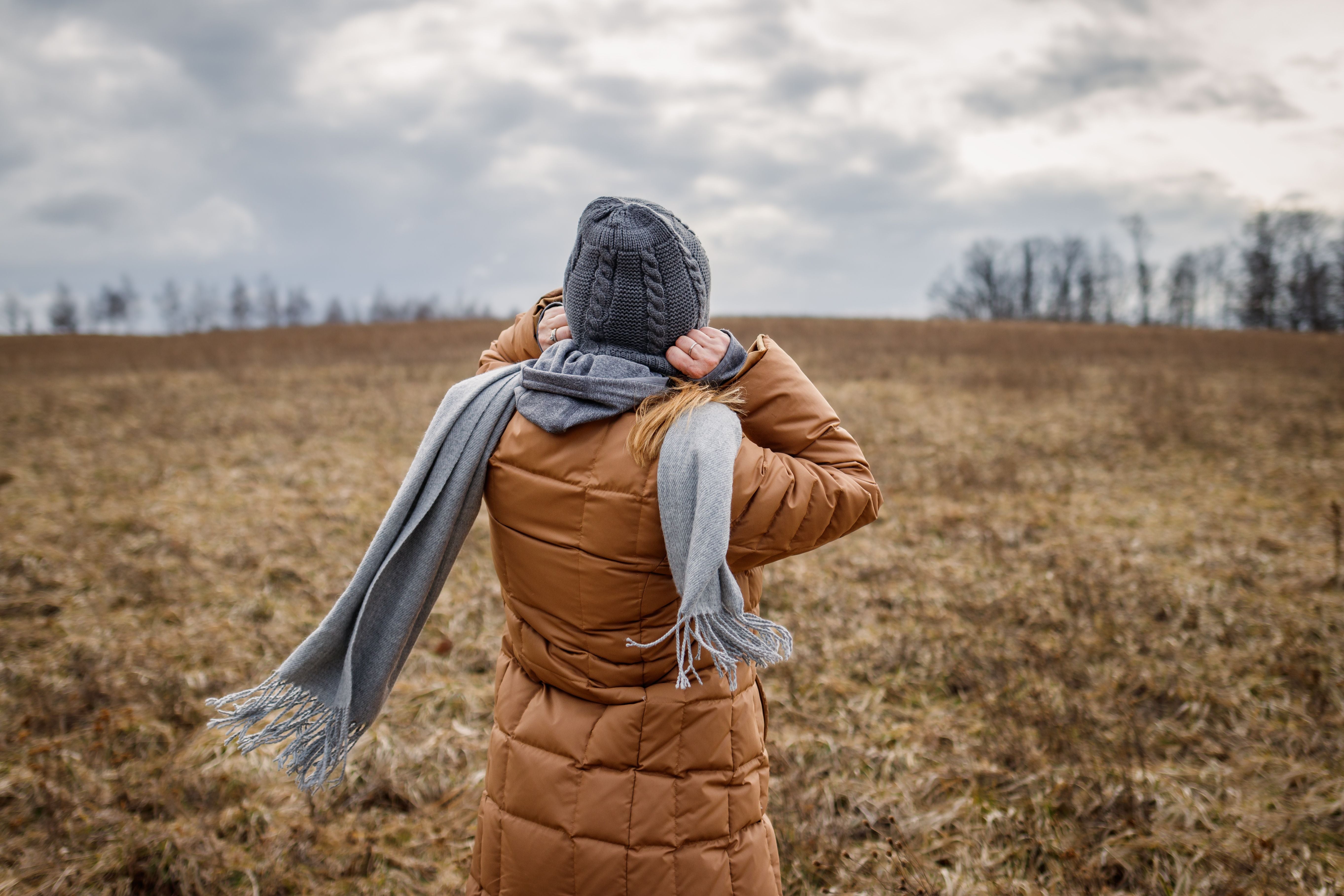Frau mit Mantel, Schal und Mütze steht an einem windigen, wolkigen Tag auf einer Wiese.