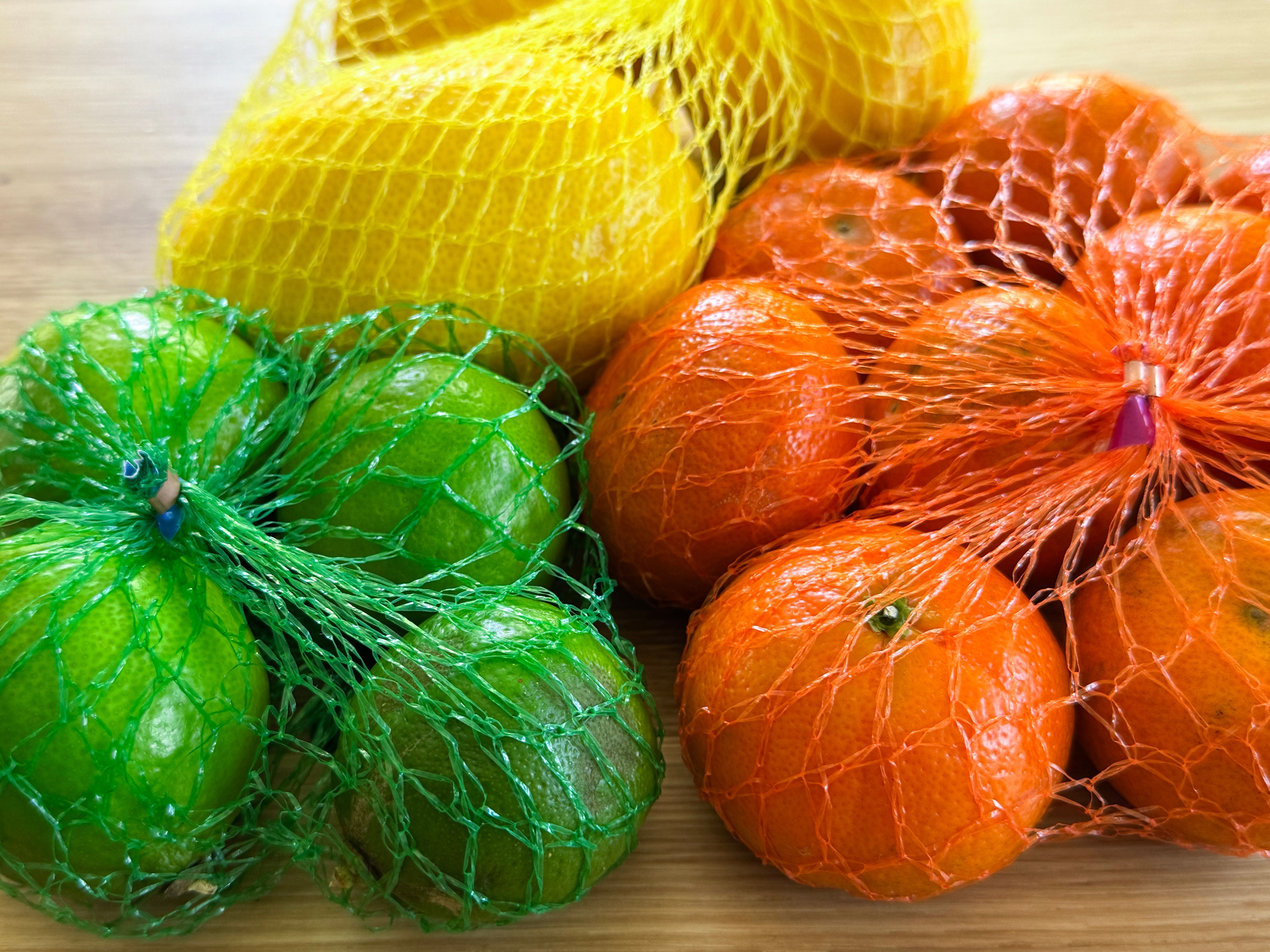 Drei Netztüten mit Limetten, Mandarinen und Zitronen auf einer Holzoberfläche.