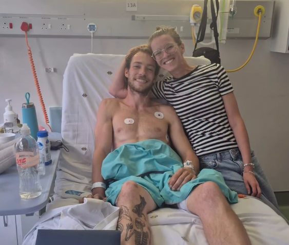 Ein Mann liegt mit Verbänden im Krankenhausbett, eine Frau sitzt lächelnd neben ihm.