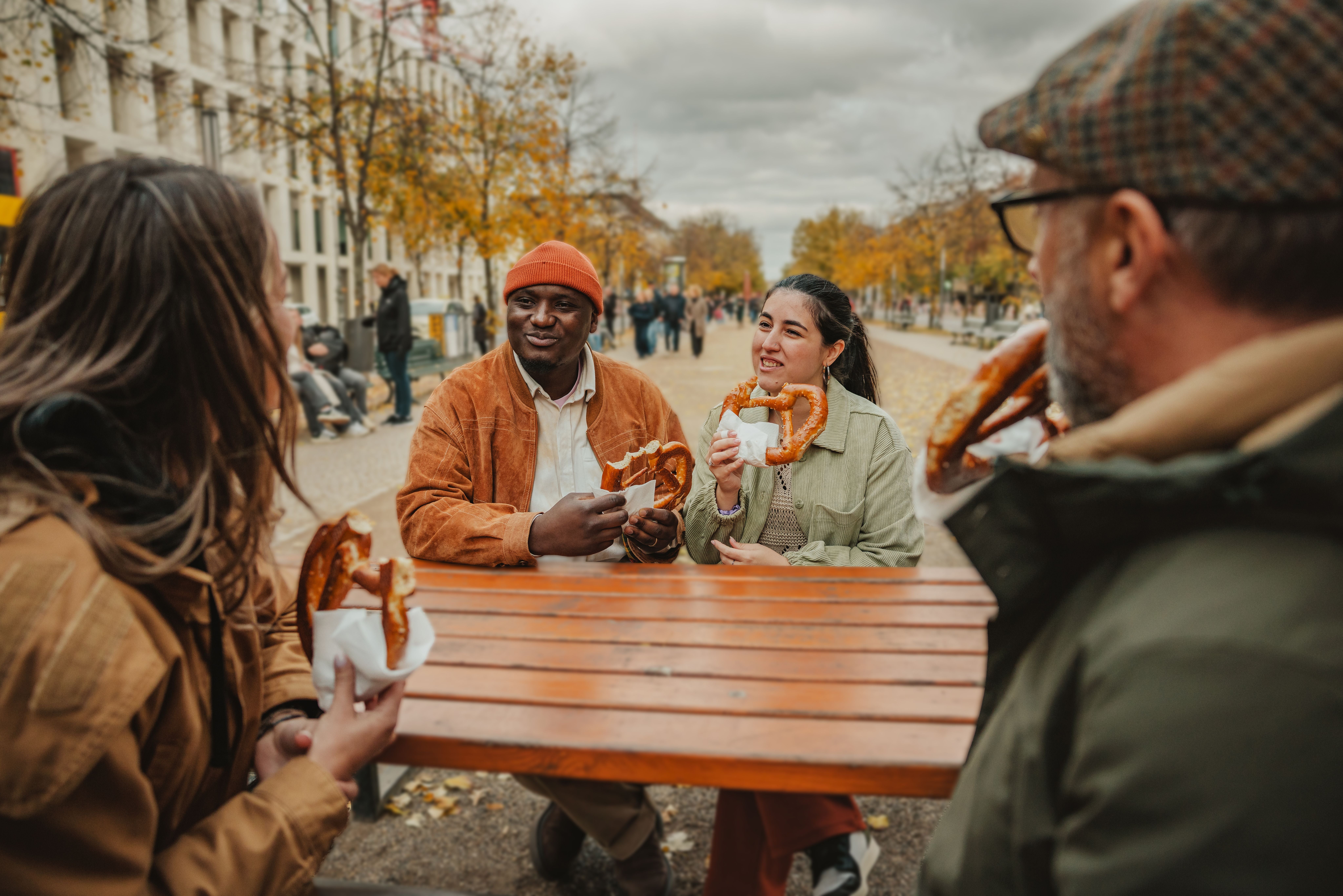 Vier Freunde sitzen draußen an einem Tisch in Berlin und essen lachend Brezeln zusammen.