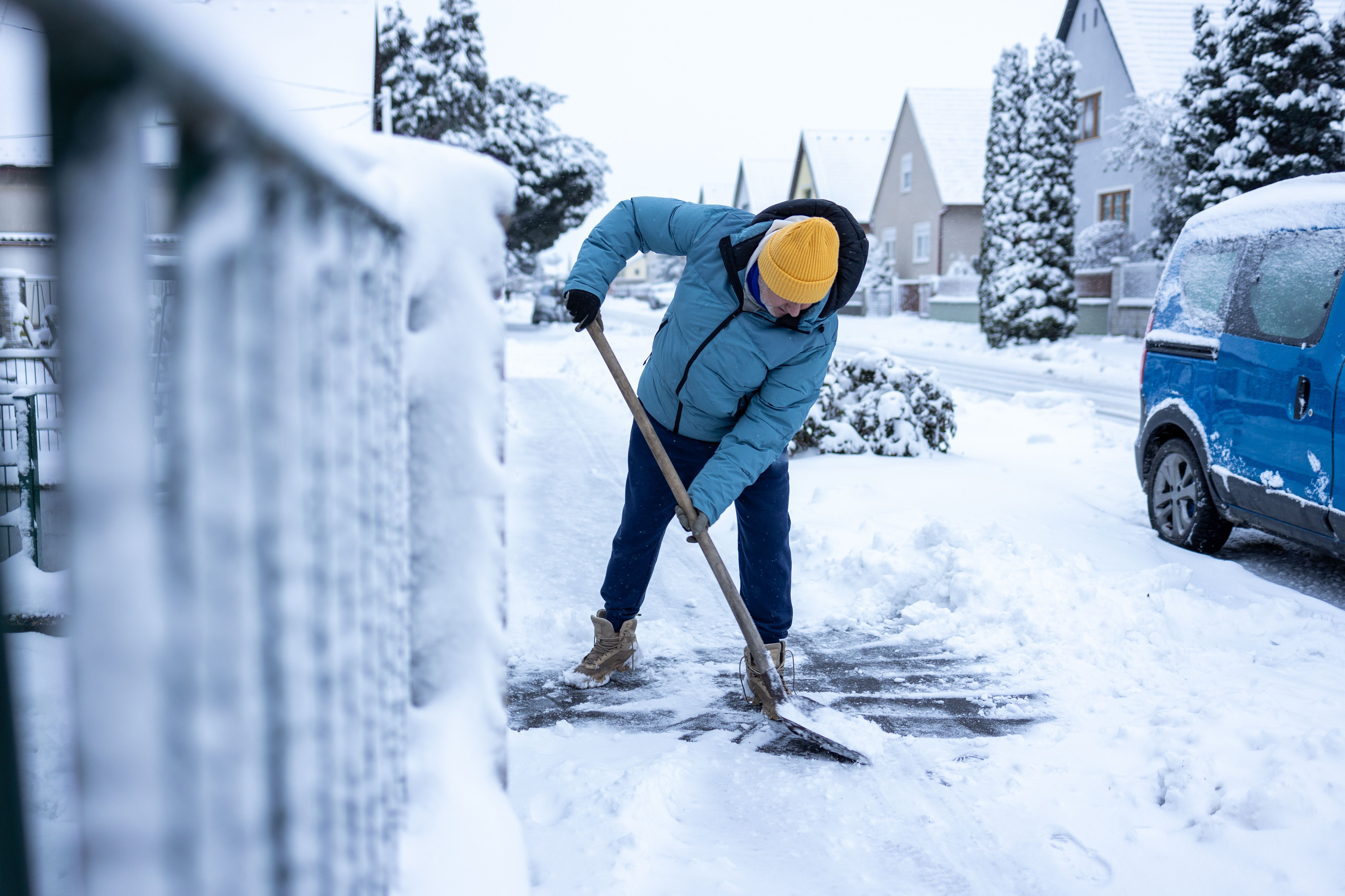 Älterer Mann schaufelt Schnee von einem Gehweg in einem verschneiten Wohngebiet.