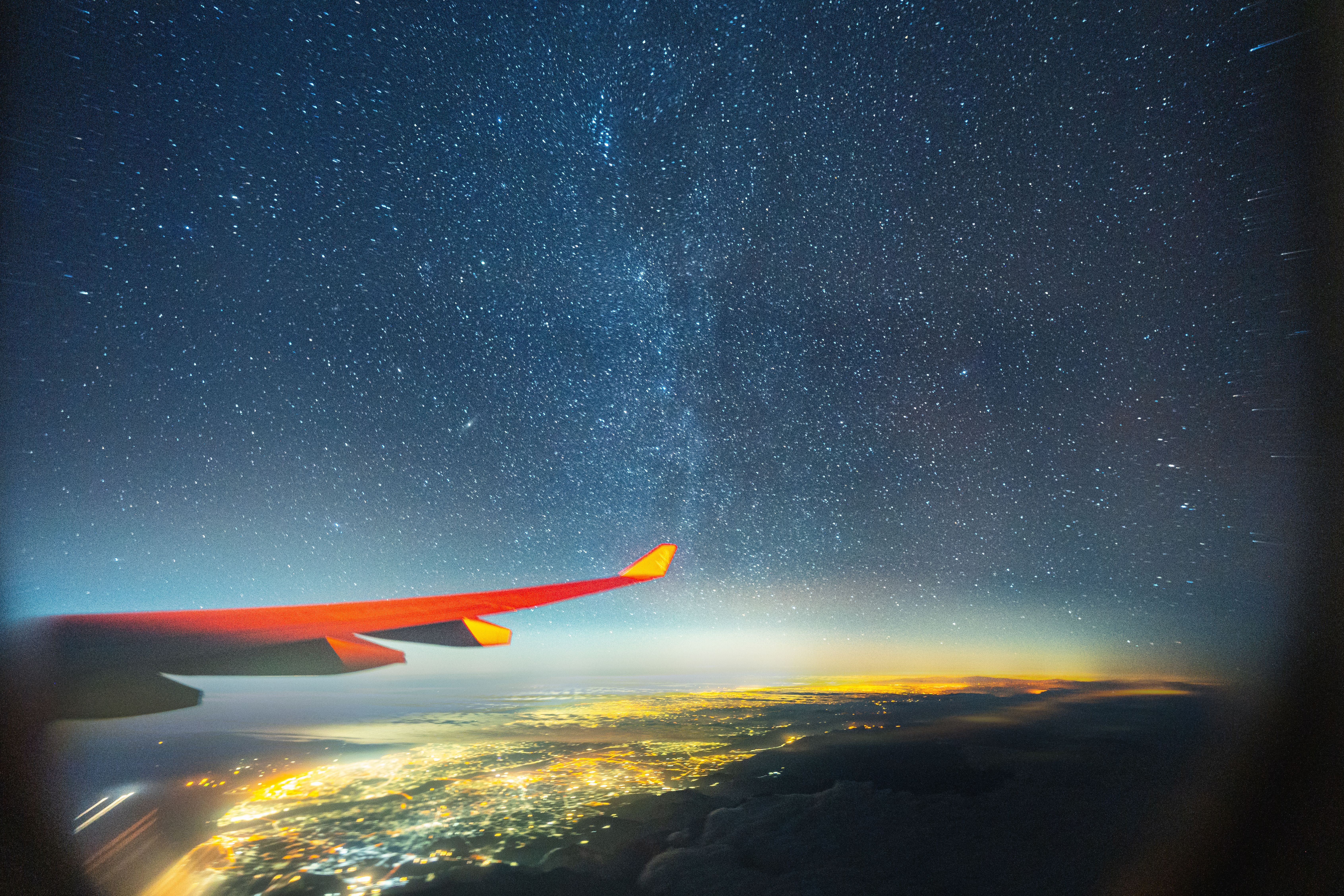 Flugzeugflügel über beleuchteter Stadt bei Nacht mit sichtbarer Milchstraße am Sternenhimmel.