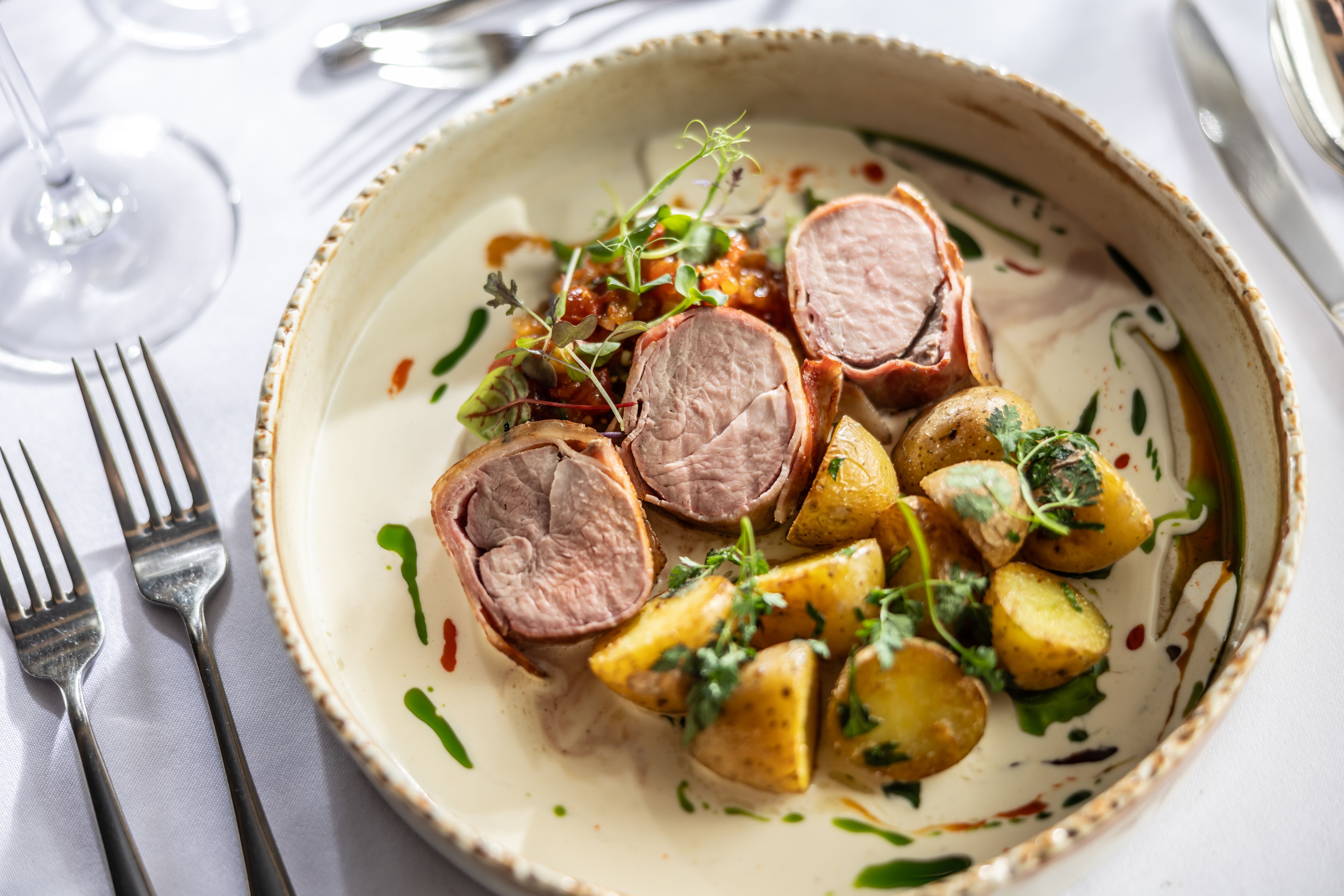 Schweinefilet im Speckmantel mit Bratkartoffeln, Cremesauce, Kräutern und Öltupfern modern angerichtet.