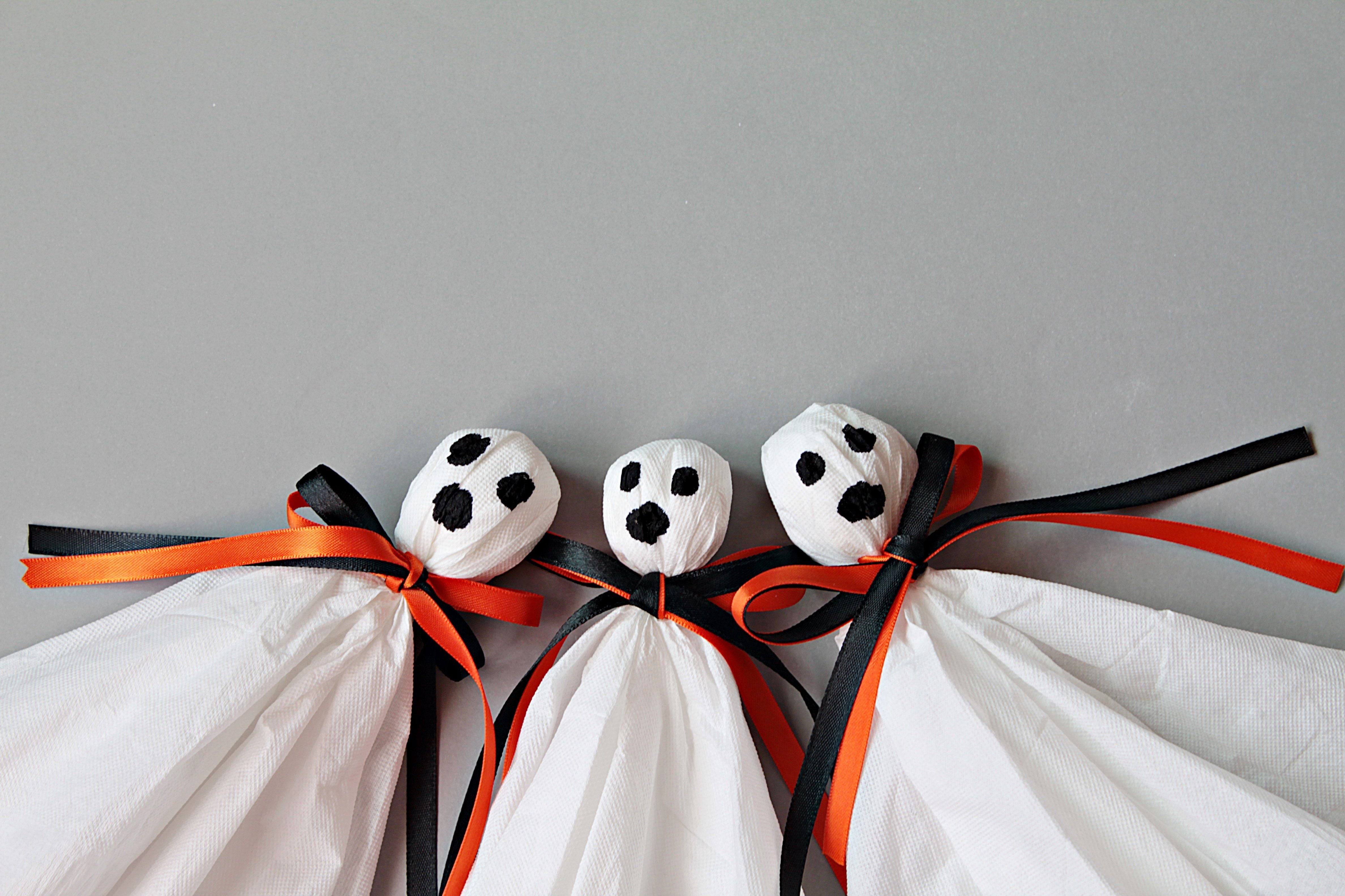 Drei DIY-Geister aus Papiertüchern mit schwarzen und orangefarbenen Bändern auf grauem Hintergrund.