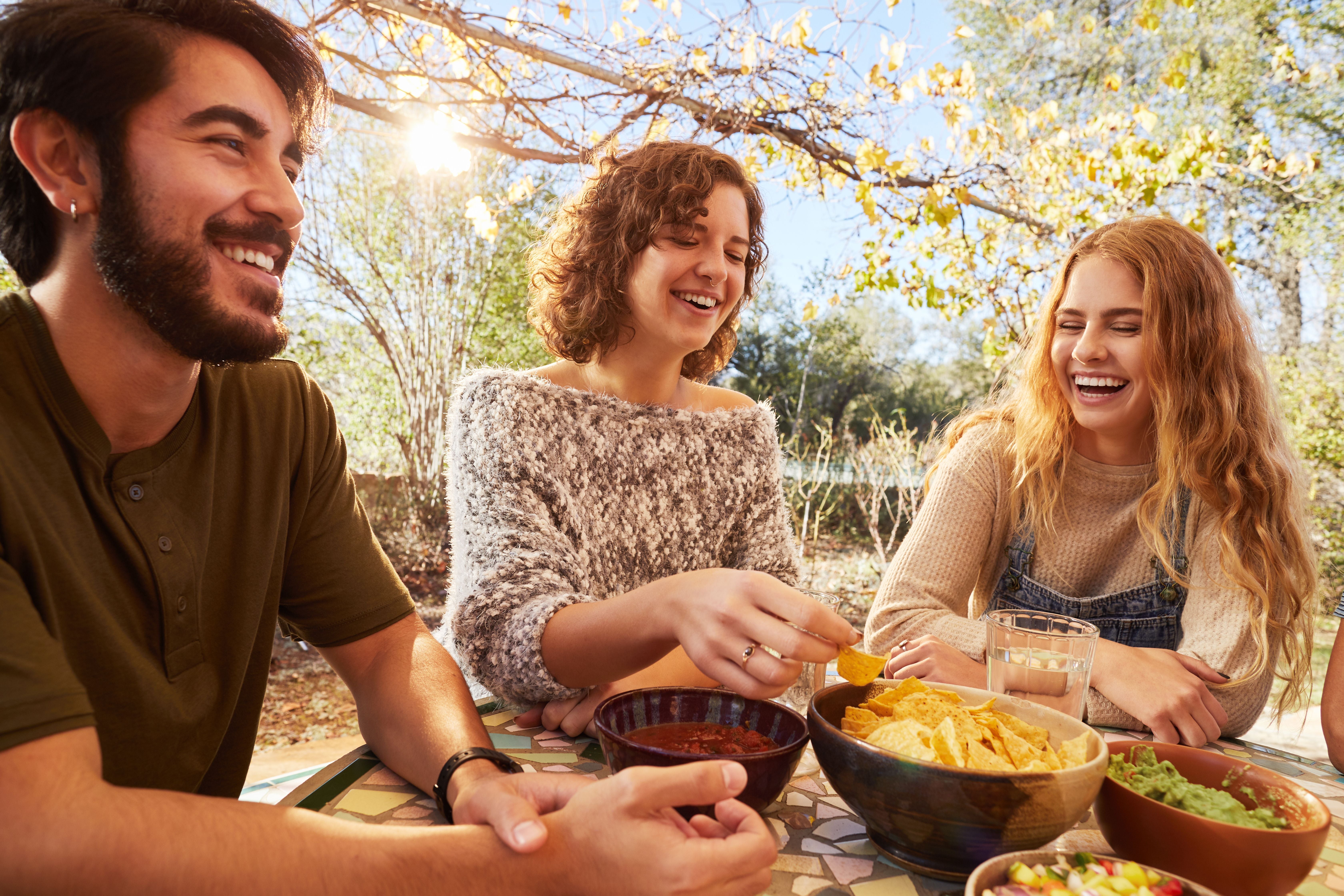Drei junge Freunde lachen und essen Snacks zusammen auf einer sonnigen Terrasse im Freien.