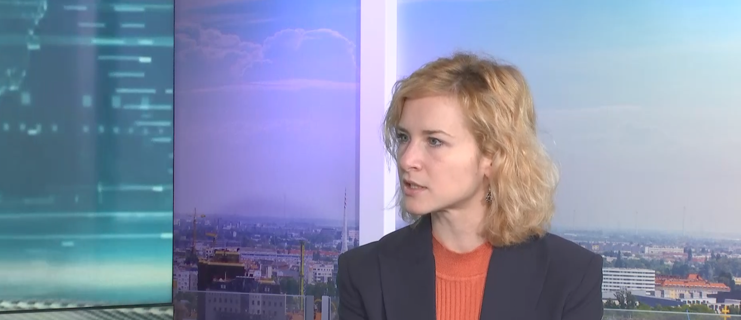Eine Frau mit blonden Haaren spricht in einem Fernsehstudio vor Stadtpanorama.