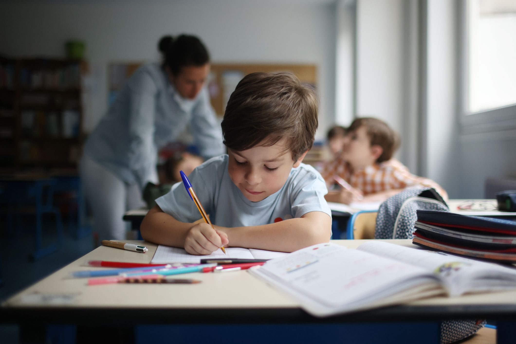 Junge sitzt an einem Schultisch und schreibt konzentriert in sein Heft, im Hintergrund Mitschüler.