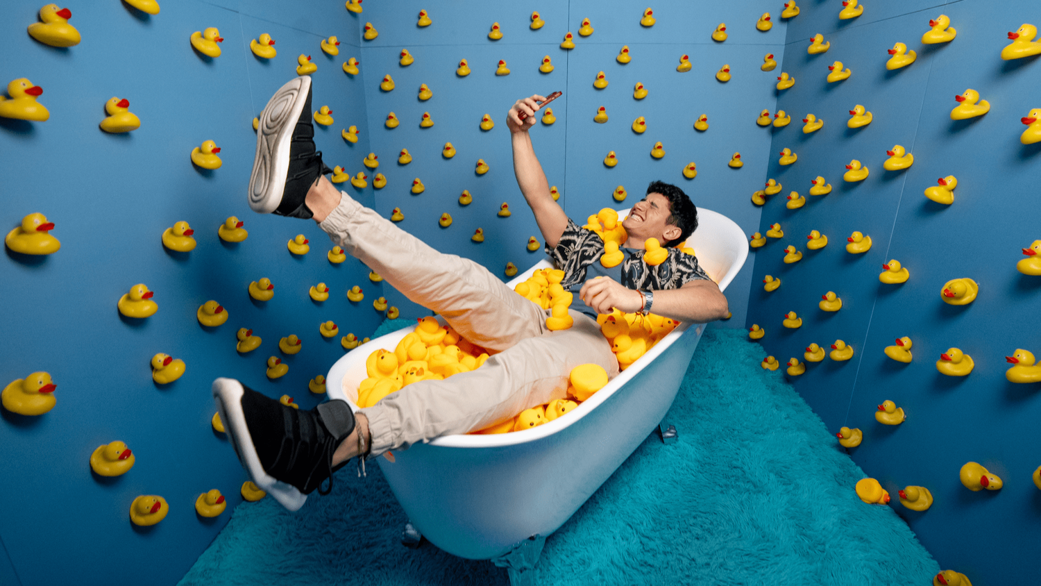 Junger Mann macht Selfie in Badewanne voller Quietscheenten, umgeben von Wänden mit Enten.