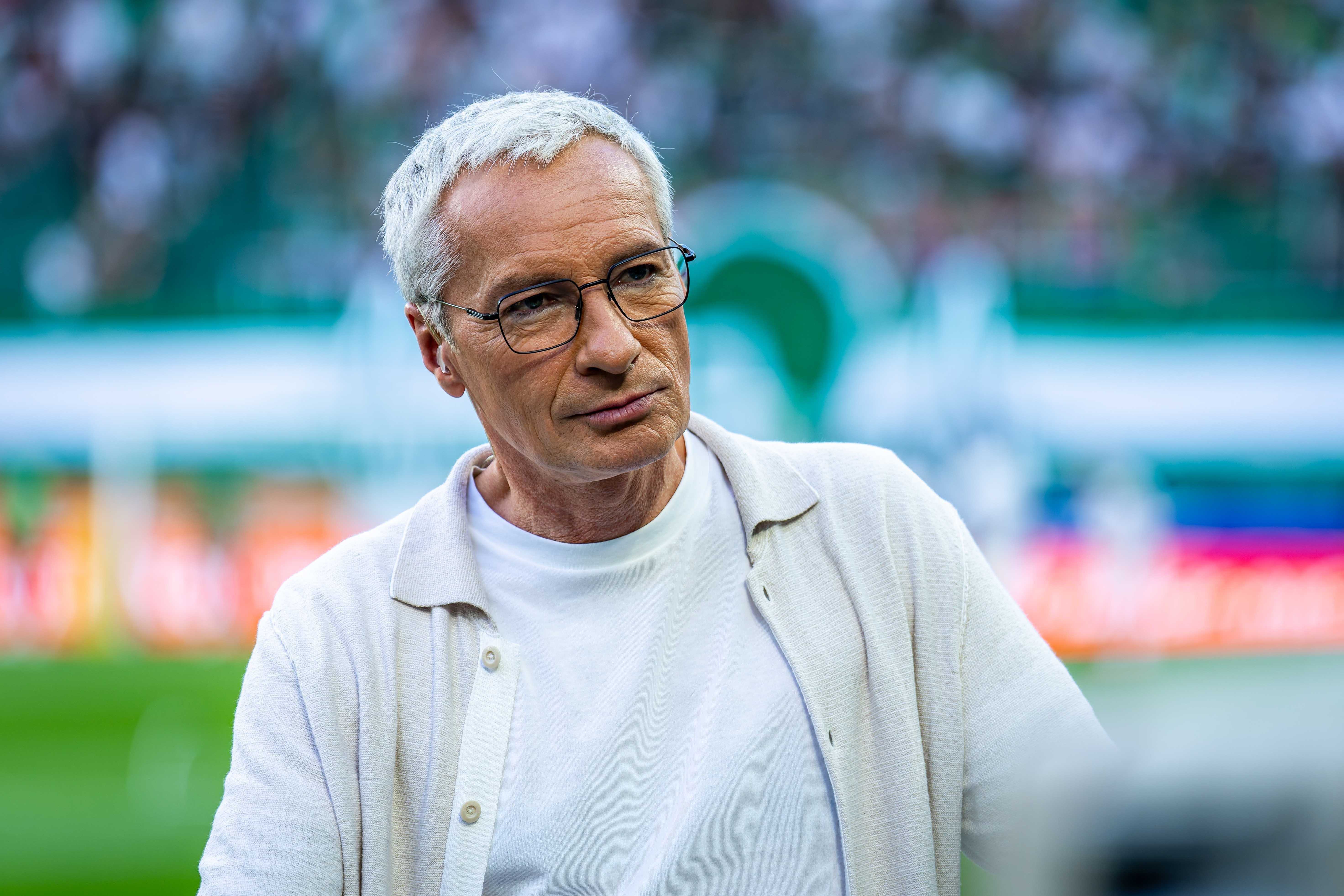 Ein älterer Mann mit Brille steht in einem Stadion und schaut nachdenklich zur Seite.