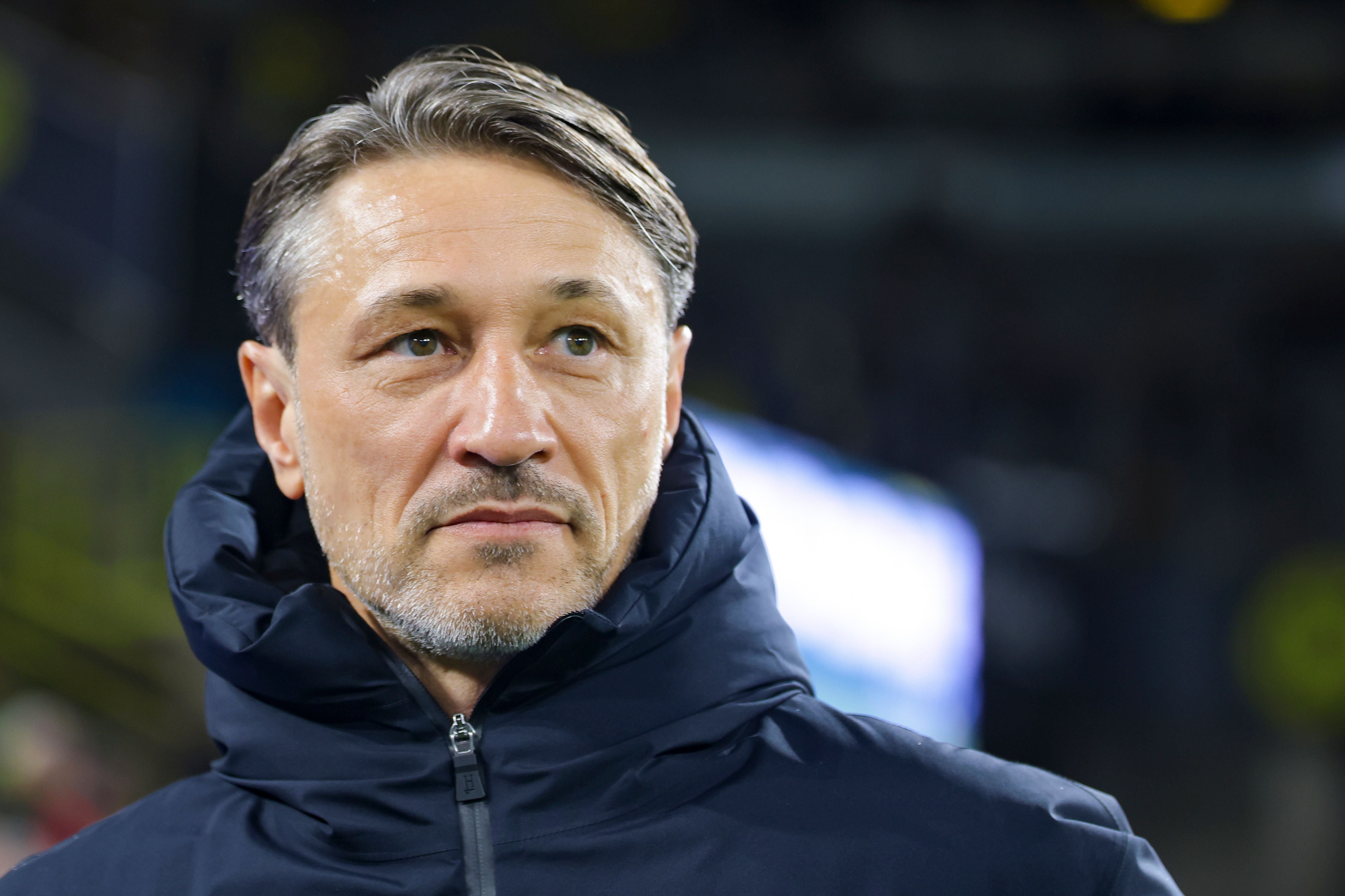 Ein Mann im dunklen Jacke blickt nach links, unscharfer Hintergrund in einem Stadion.
