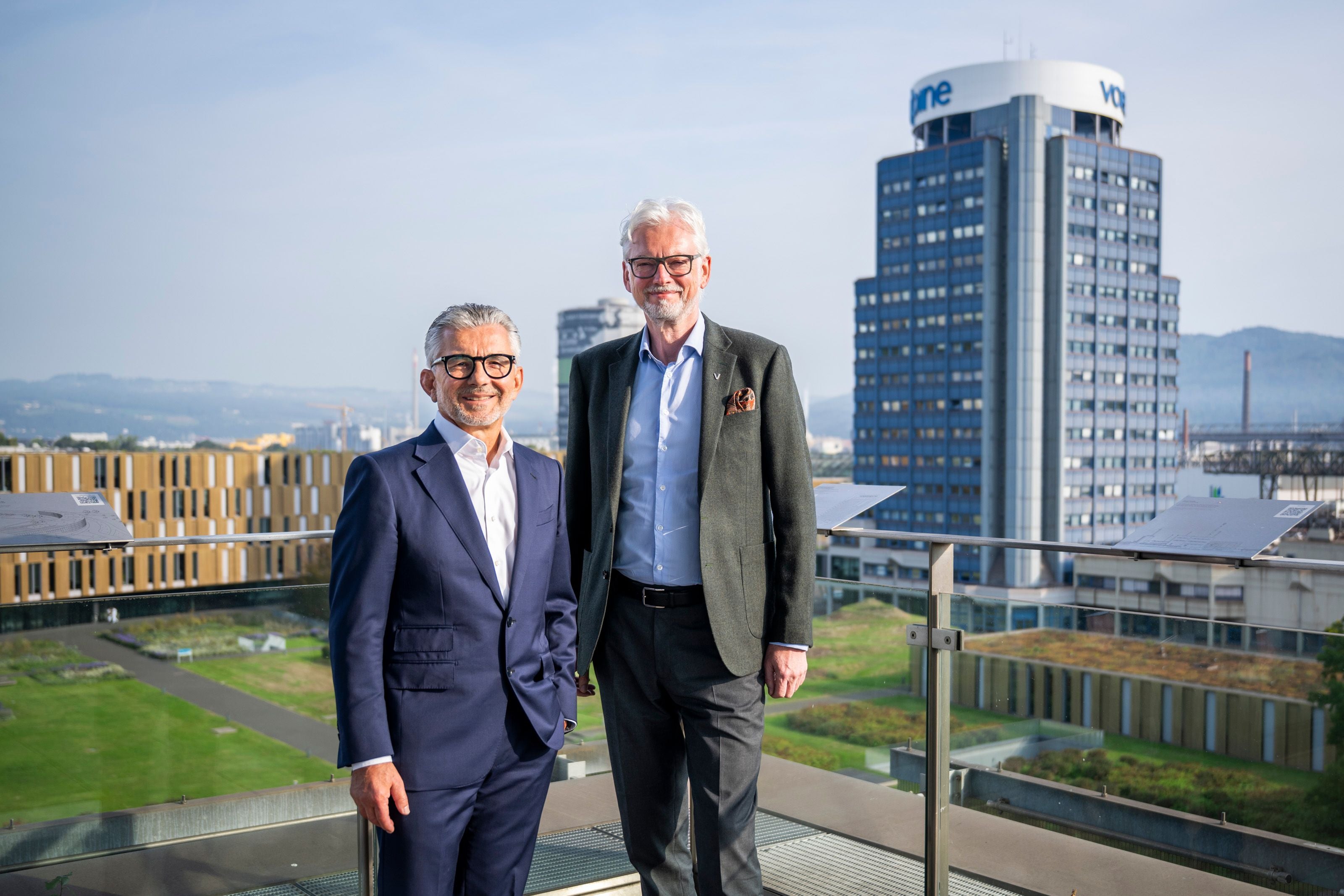 Zwei Männer im Anzug stehen auf einer Terrasse vor modernen Bürogebäuden und blicken in die Kamera.