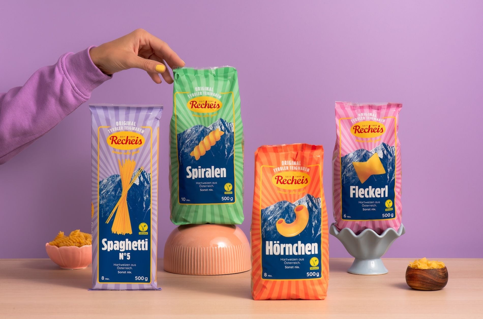 Vier Recheis Nudelpackungen (Spaghetti, Spiralen, Hörnchen, Fleckerl) vor lila Hintergrund.
