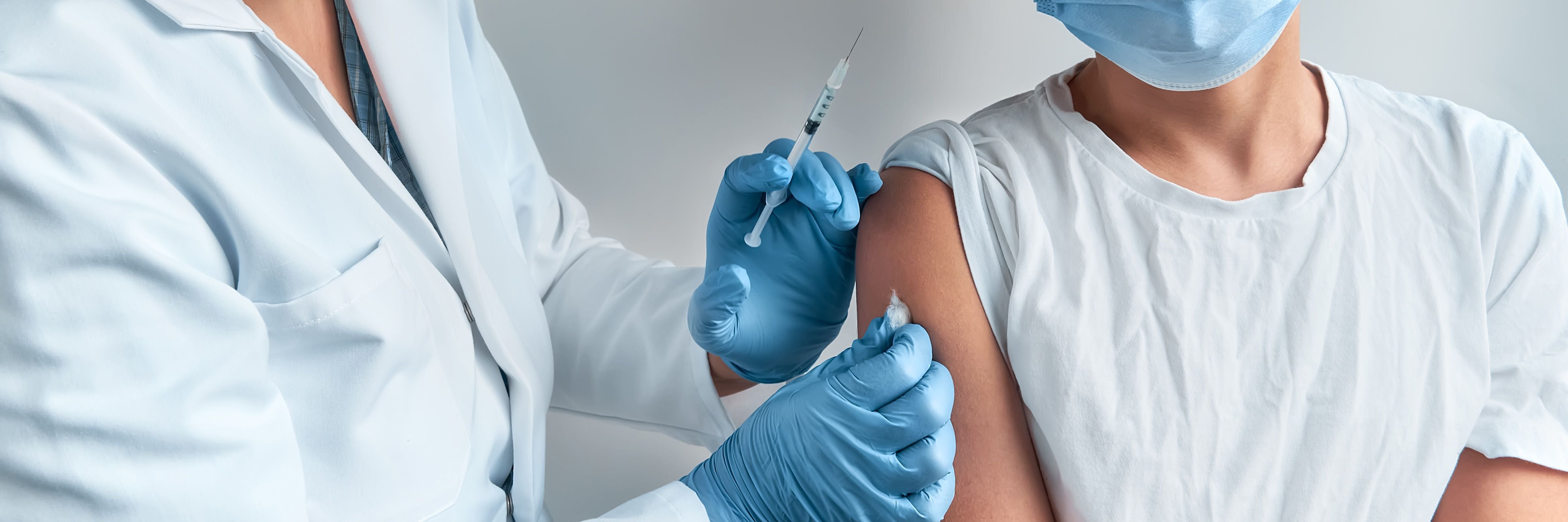 Ärztin impft Patientin in den Oberarm, beide tragen Schutzkleidung und Masken.