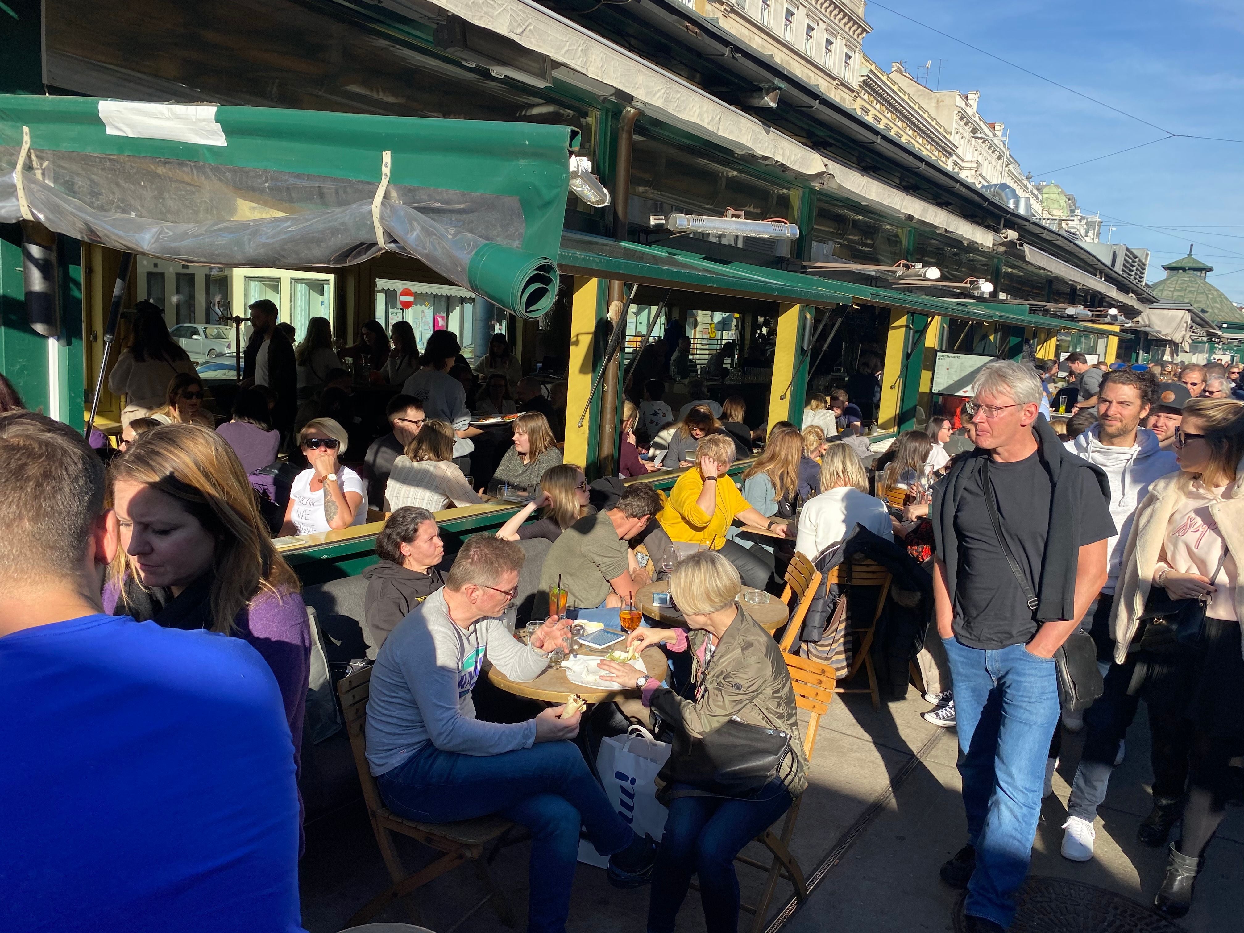 Viele Menschen sitzen und stehen an einem belebten Straßencafé bei Sonnenschein.