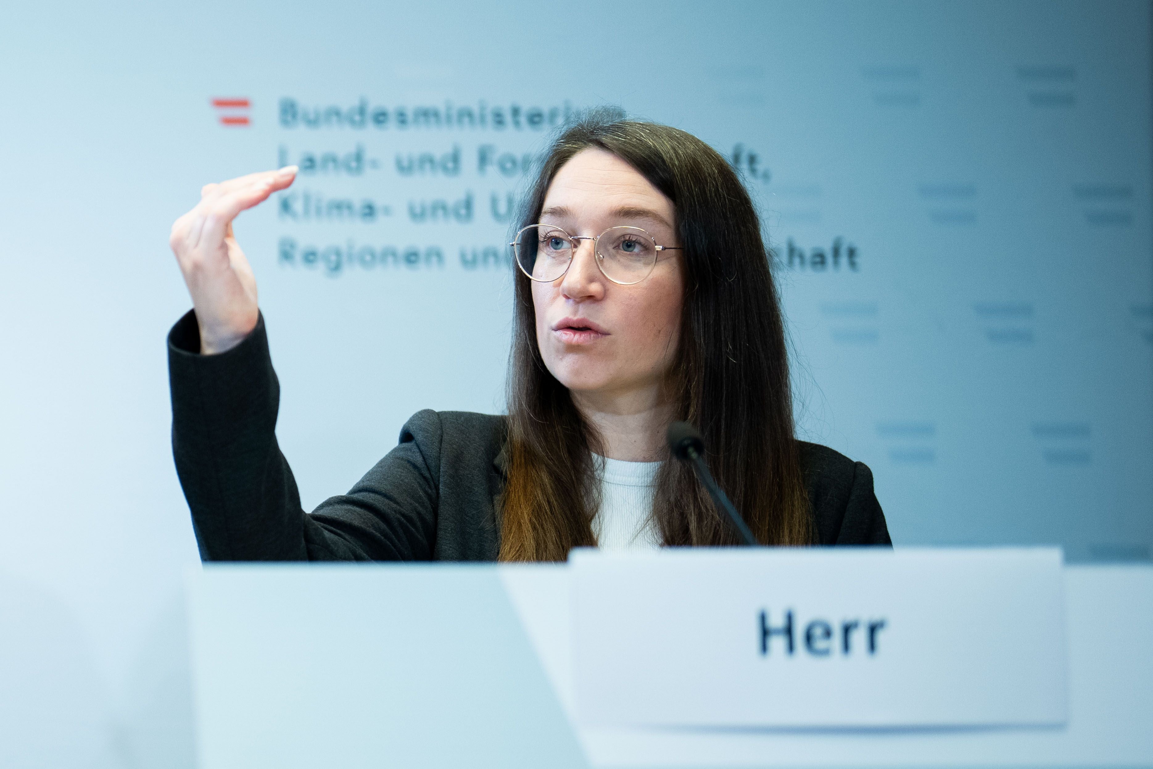 Frau spricht während einer Pressekonferenz am Rednerpult mit Mikrofon und Namensschild „Herr“.