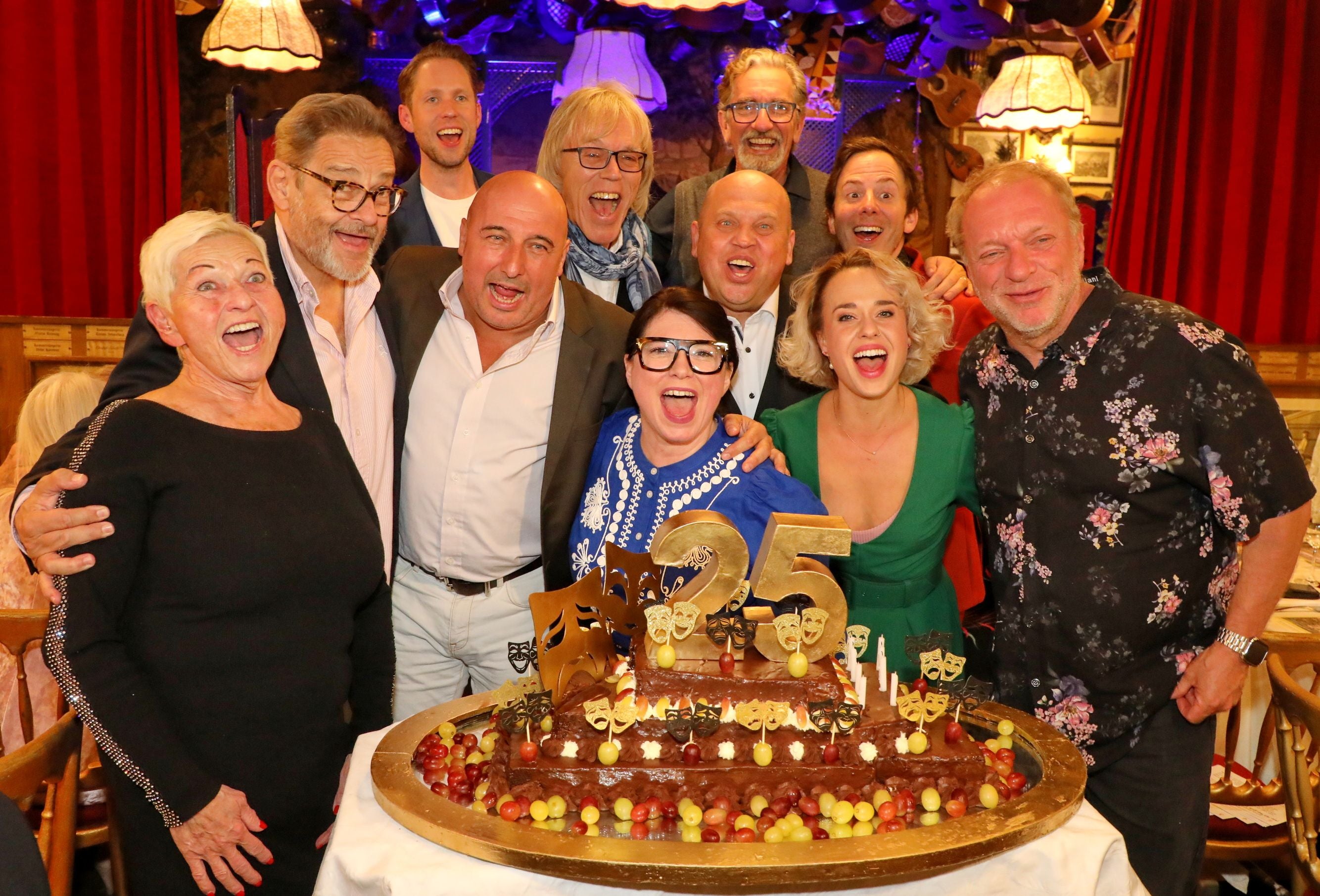 Eine fröhliche Gruppe feiert mit einer großen „25“-Jubiläumstorte im festlich geschmückten Raum.