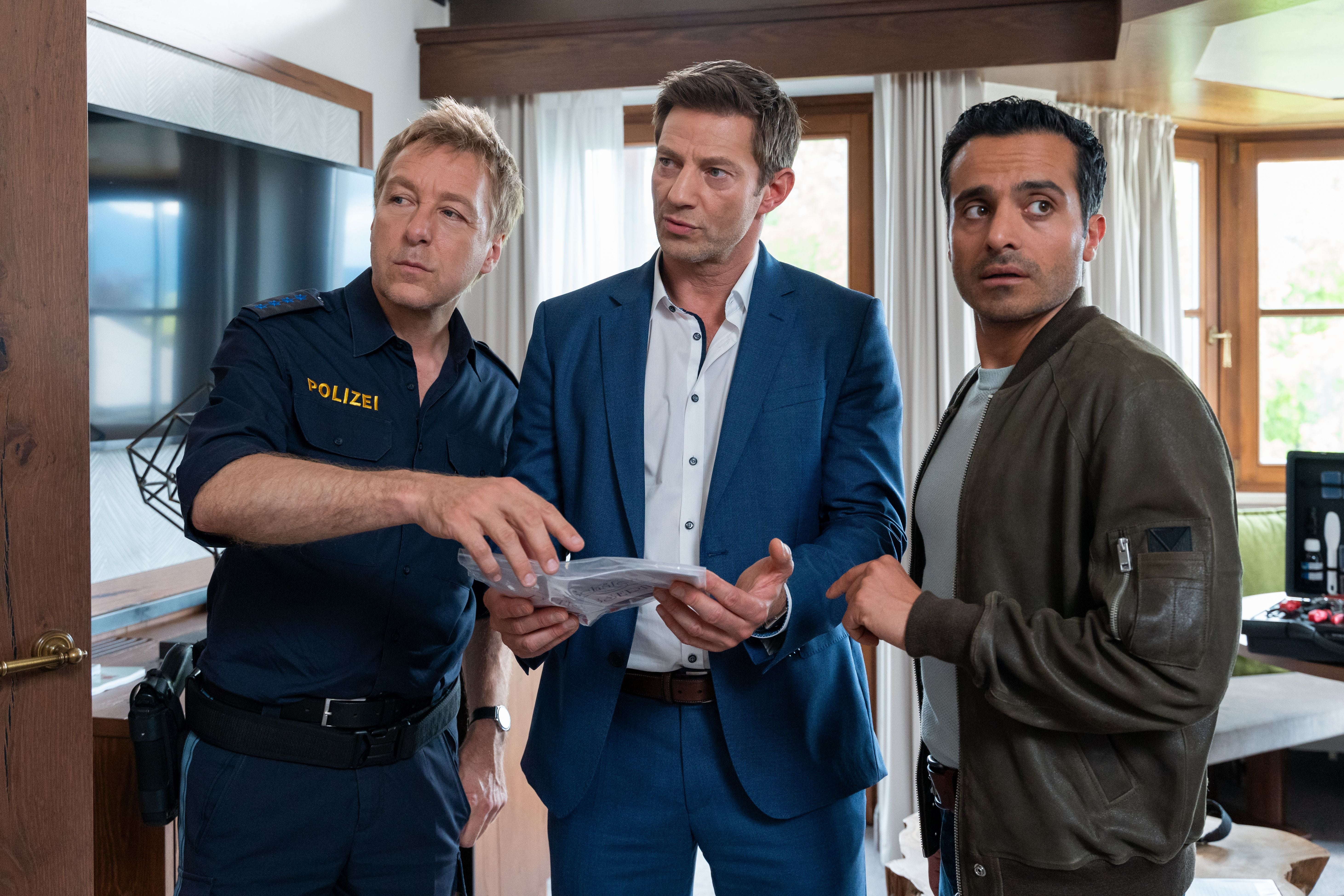 Drei Männer, einer in Polizeiuniform, betrachten gemeinsam ein Dokument in einem Wohnzimmer.