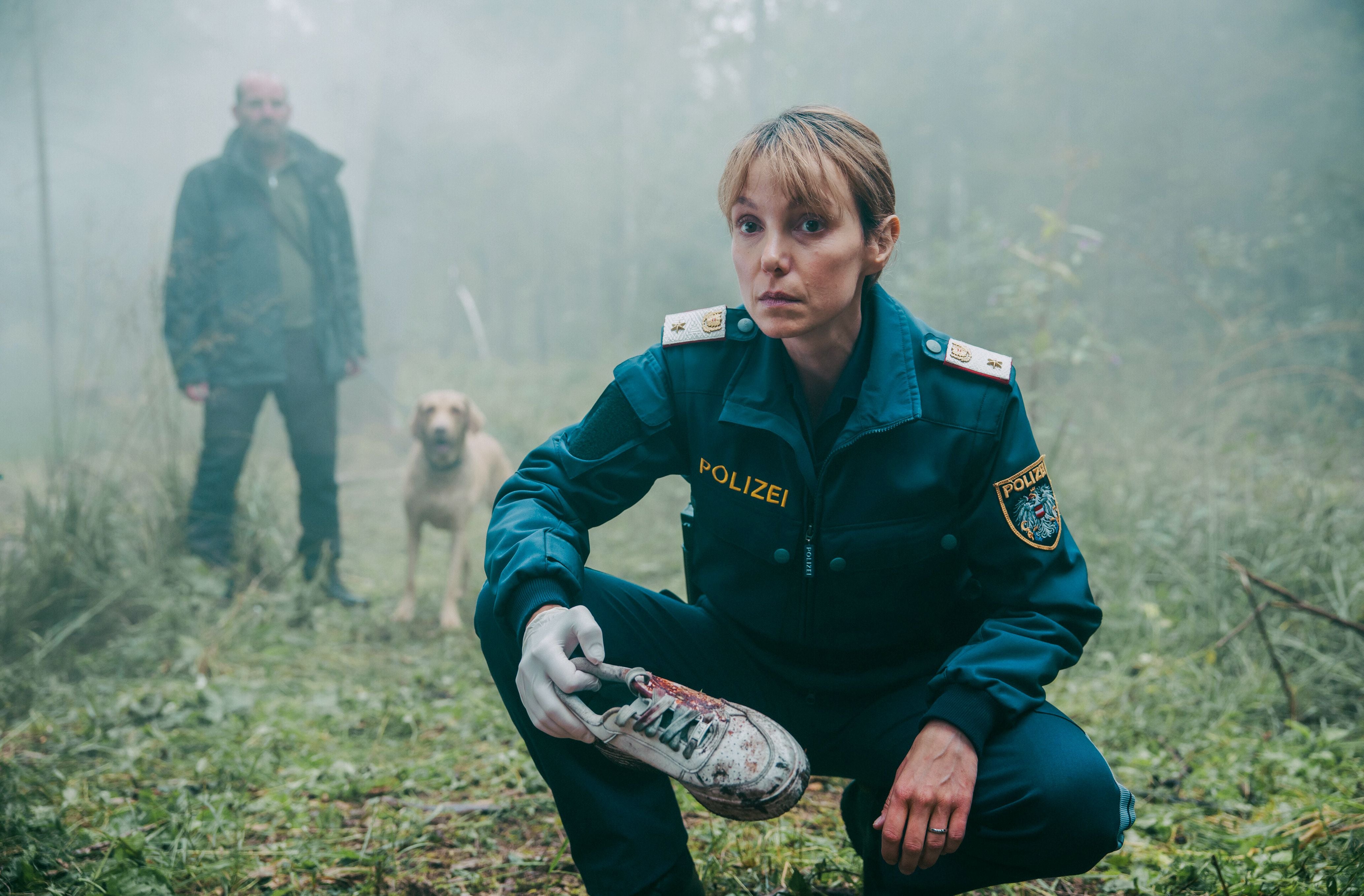 Polizistin hält blutverschmierten Turnschuh, im Hintergrund ein Mann mit Hund im nebligen Moor.