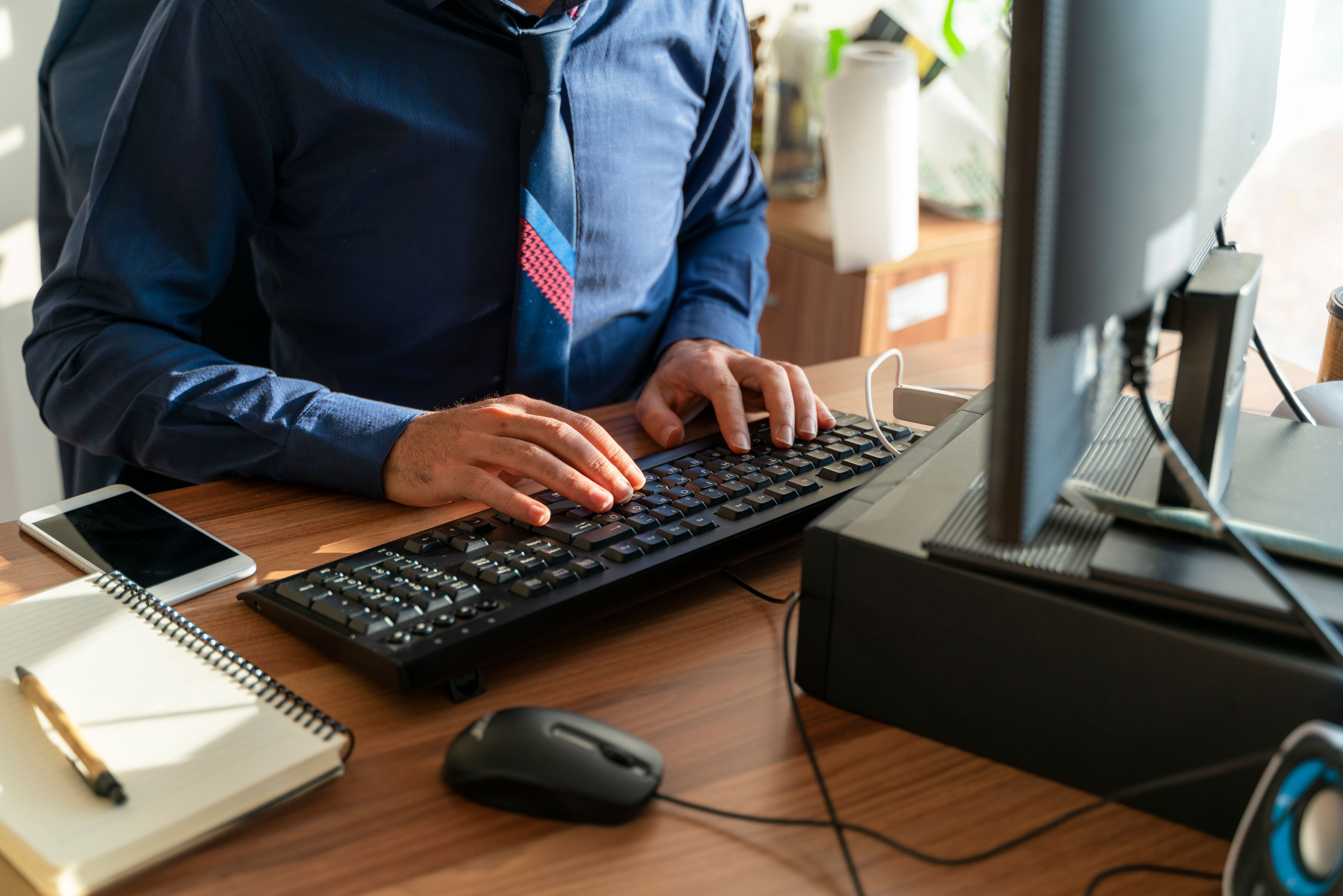 Ein Mann tippt in einem Büro am Computer auf einer Tastatur, neben Notizbuch und Handy.