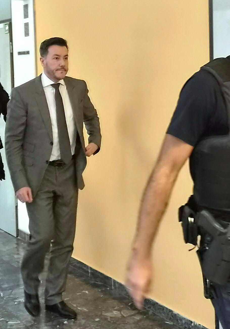 Ein Mann im grauen Anzug geht durch einen Flur, im Vordergrund ein Polizist mit Weste.