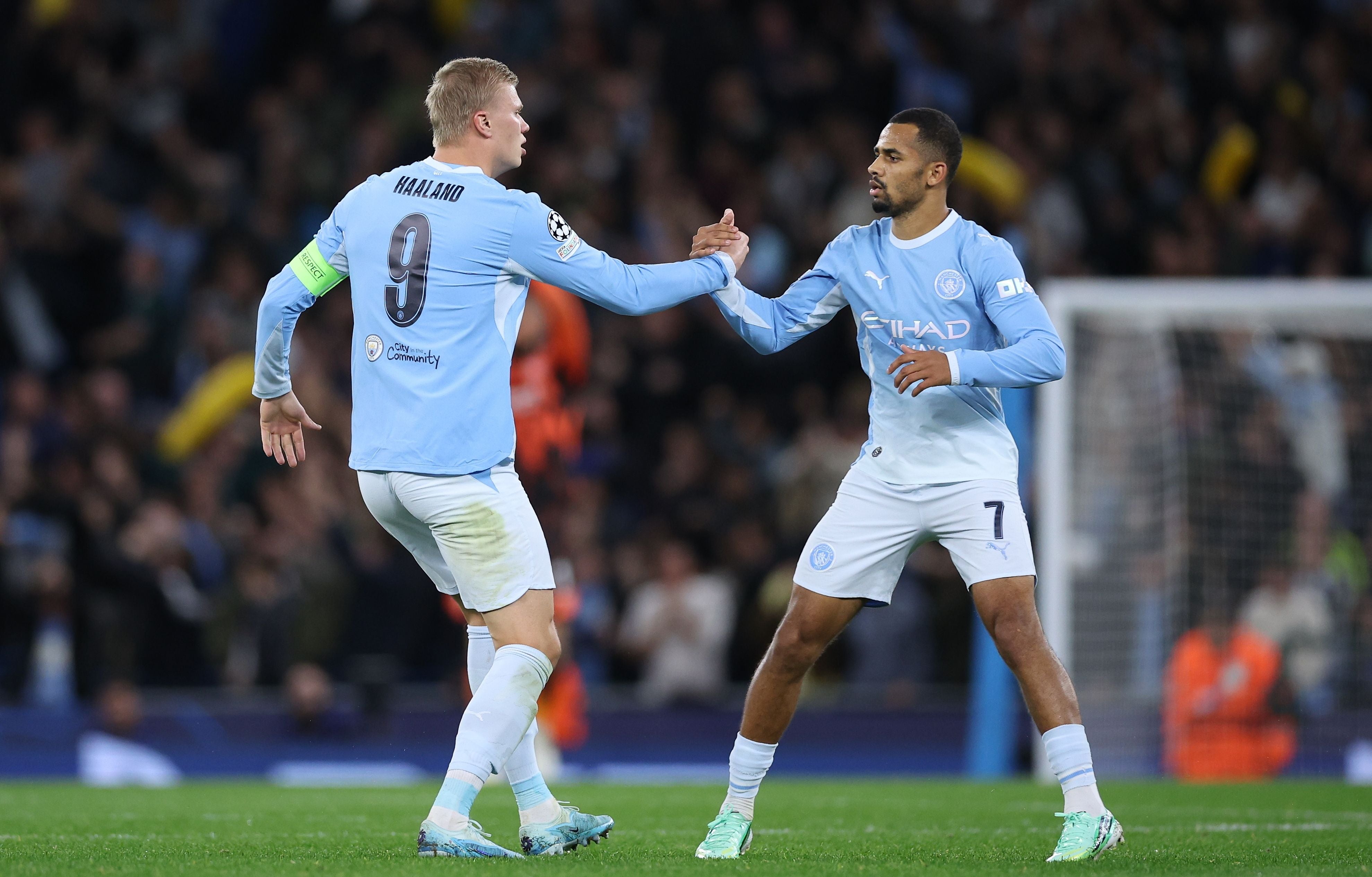 Erling Haaland mit der Rückennummer 9 und Kapitänsbinde klatscht auf dem Spielfeld im hellblauen Trikot von Manchester City mit seinem Teamkollegen Gabriel Jesus ab.
