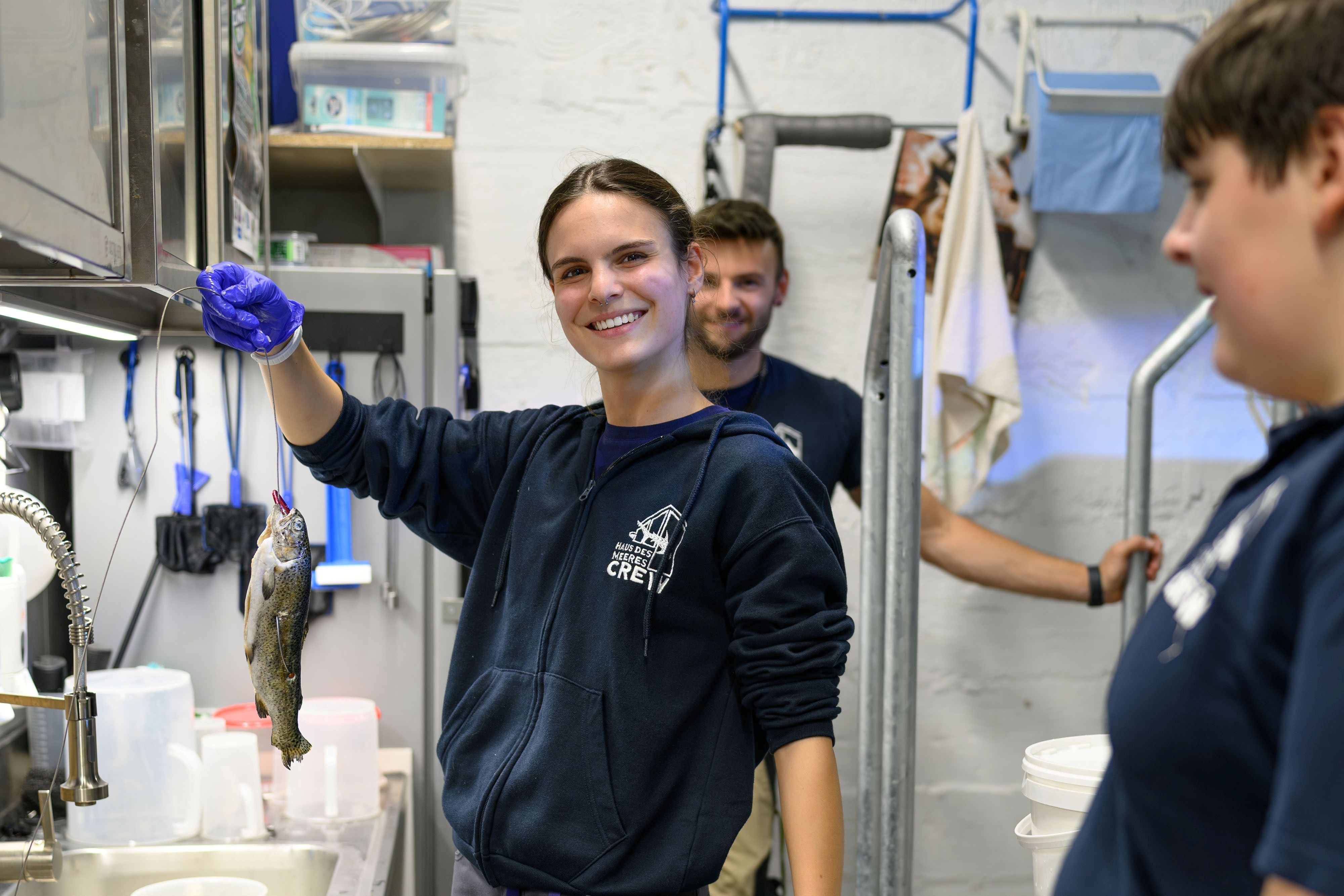 Eine Frau hält einen Fisch in einem Labor, während zwei weitere Personen zuschauen.